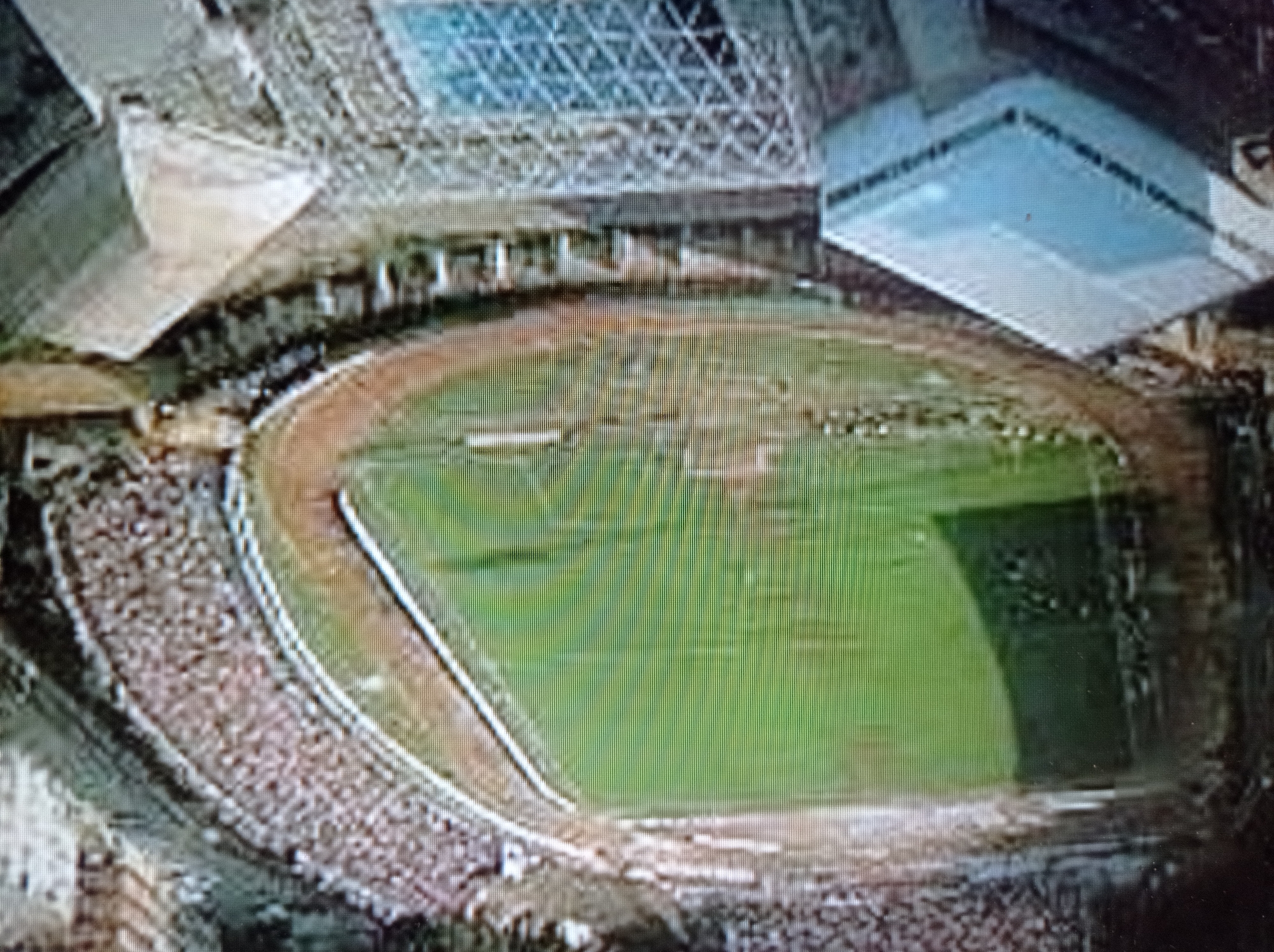This is an aerial, low-resolution image of a stadium in Mukachevo, Ukraine. The stadium features a green field, possibly a soccer pitch, surrounded by a running track. Bleachers filled with spectators line a portion of the stadium. In the foreground, there are modern structures, including a building with a light-colored roof and a large glass-paneled roof or skylight. The surrounding environment appears to be urban or semi-urban, with trees and other buildings visible in the distance. The image quality is grainy, suggesting it was taken with a low-resolution camera or has been compressed.