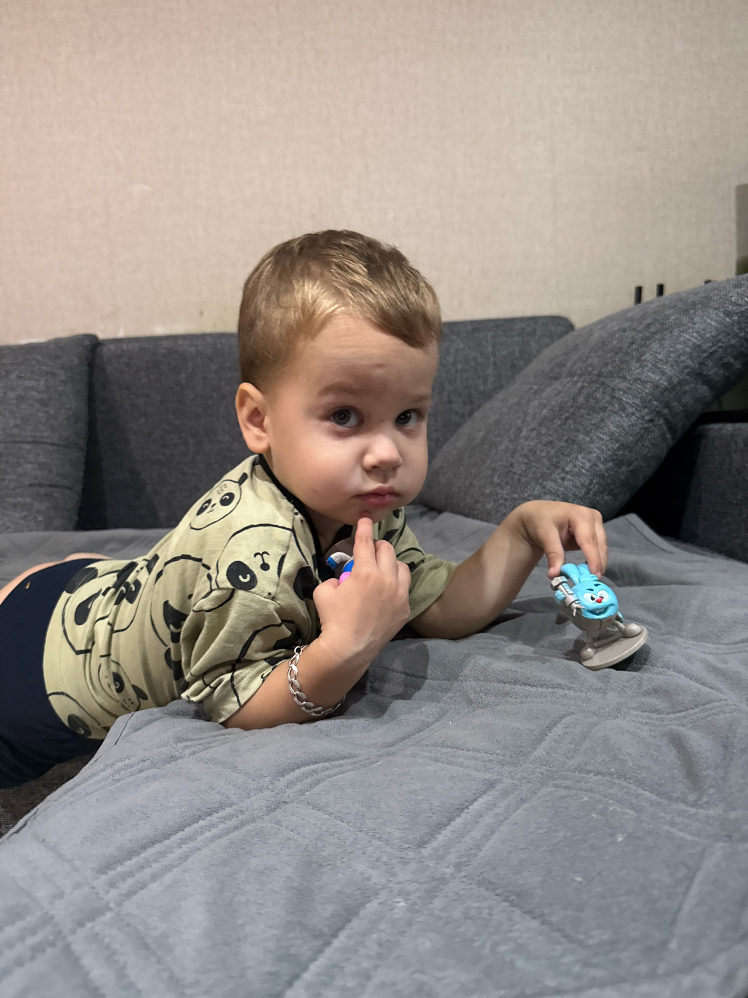 A young boy with blond hair and blue eyes is lying on a grey quilted blanket on a couch. He is wearing a light green shirt with a panda pattern and dark blue shorts. His left hand is resting on his chin, and his right hand is holding a small blue toy figure. The boy is looking directly at the camera with a thoughtful expression. Behind him, a dark grey couch is visible, with a textured wall in the background. The overall impression is a candid indoor portrait of a child at play. This image is not located in Rusanovo, Russia.