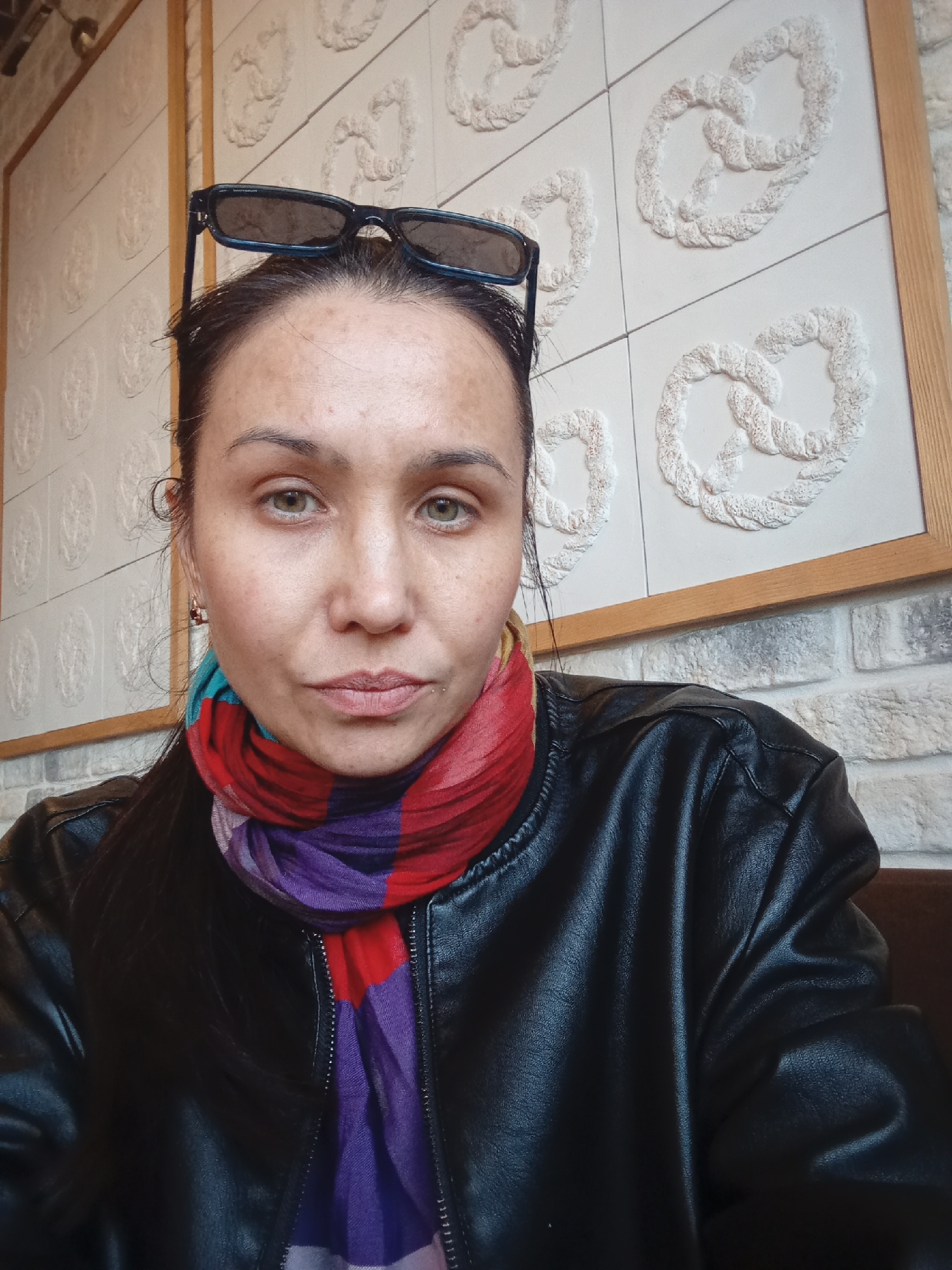 A medium close-up shows a woman wearing a black leather jacket and a colorful scarf. The scarf features red, purple, and blue colors. Dark-rimmed sunglasses are perched on top of her head. She has green eyes and dark hair pulled back. The background includes a wall decorated with a pattern of pretzels in white relief, framed by a wooden border. The lower portion of the wall appears to be made of white bricks. The photo was likely taken indoors. There is no specific evidence to confirm that the photo was taken in Pervomaysky, Russia.