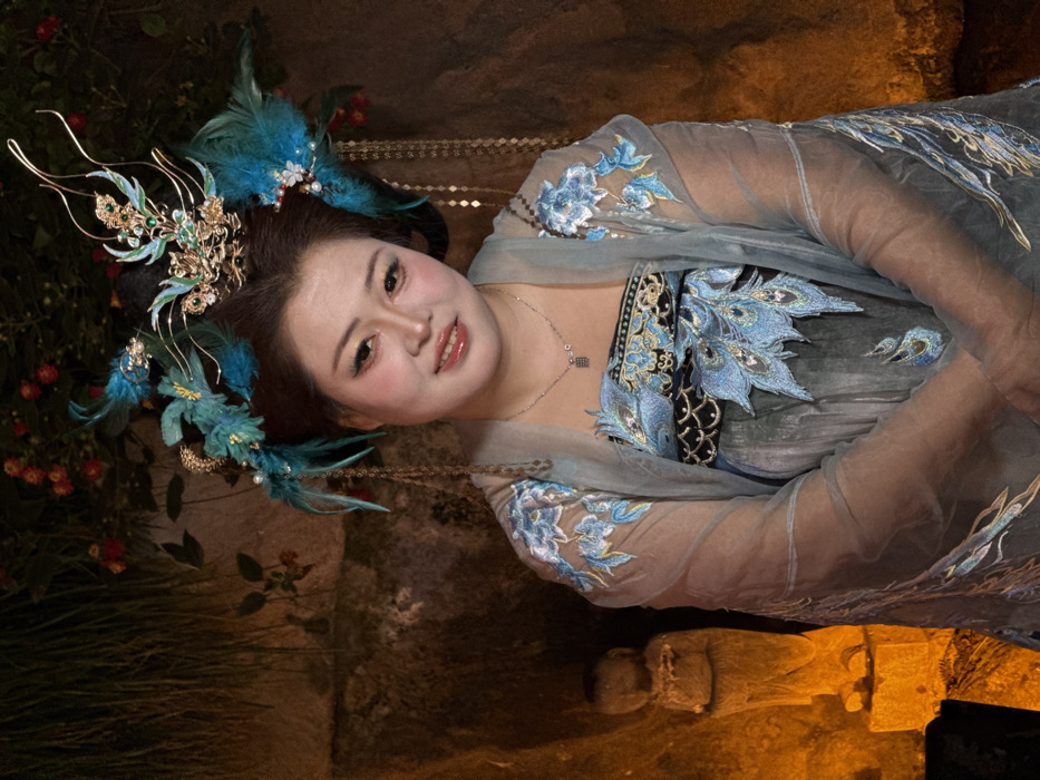 The image displays a posed portrait of a young East Asian woman, smiling softly and looking directly at the viewer. The photograph is taken in low light conditions, likely at night, and is oriented at a 90-degree angle, making it appear as if the subject is reclining.
The woman has dark hair styled in an elaborate updo, adorned with a magnificent headpiece. The headdress features intricate gold-colored metalwork shaped into floral and flowing patterns, accented with green and blue enamel or gems. Large, vibrant teal-blue feathers are strategically placed within the hairstyle, adding volume and color. Small pearl-like beads and delicate gold-colored chains dangle from the headdress, gracefully extending towards her shoulders. Her face is made up with blush, winged eyeliner, and a reddish lip gloss, enhancing her pleasant expression.
She is dressed in an ornate, multi-layered costume reminiscent of traditional East Asian attire. The outermost layer is a sheer, light blue-grey fabric, possibly chiffon or organza, featuring delicate light blue floral embroidery on the sleeves and upper body. Beneath this, her inner garment or bodice is visible, characterized by intricate black, gold, and light blue embroidery, with prominent peacock-feather-like motifs on the chest area. A slim silver-colored necklace with a small, square pendant rests around her neck.
The background is dark and somewhat indistinct. To her left (when viewed upright), there is dark foliage interspersed with small red berry-like fruits or flowers, suggesting a natural, possibly garden-like setting. To her right, the background is darker and textured, with a subtle warm orange glow appearing towards the bottom, possibly from an ambient light source reflecting off a stone surface. A vague, dark, and somewhat rectangular shape, possibly an ancient stone carving or part of a structure, is faintly discernible in the lower right portion of the image. The overall lighting accentuates the woman and her elaborate attire, casting a gentle glow on her face and the intricate details of her costume.