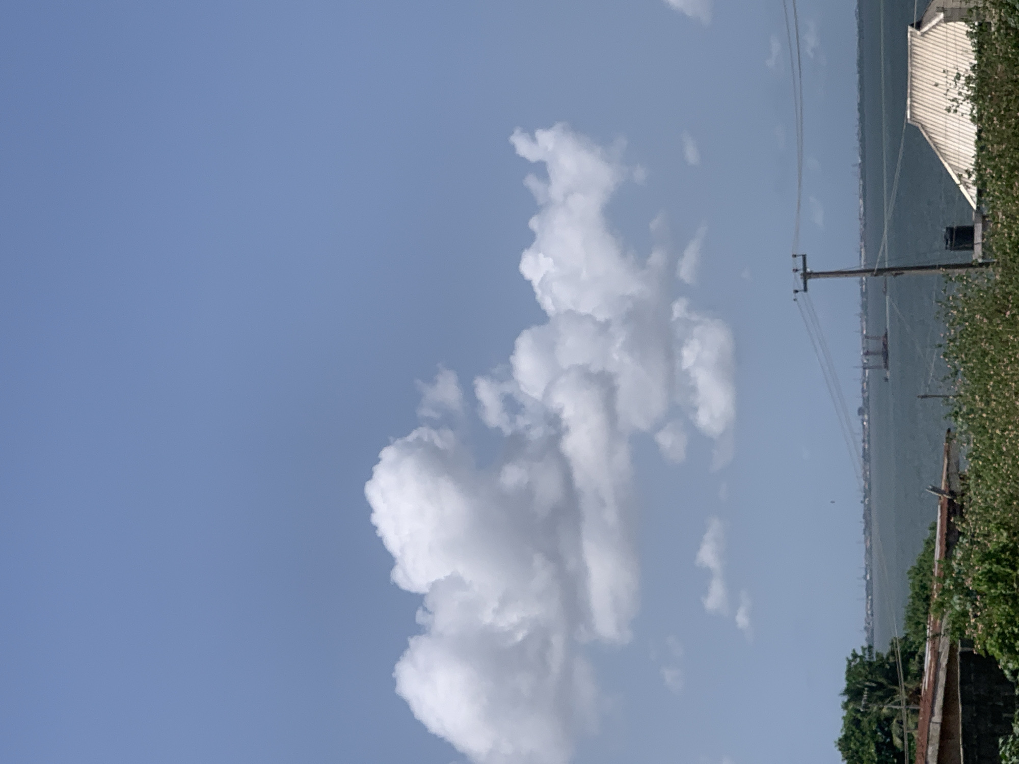 The image depicts a daytime scene in Ikorodu, Nigeria. Cumulus clouds dominate the left and center of the sky. A body of water with indistinct buildings along the opposite shore stretches across the center-right of the frame. A wooden utility pole with power lines is situated on the right side, near some foliage. Part of a building with a white roof is partially visible in the upper right corner. The image is rotated counter-clockwise.