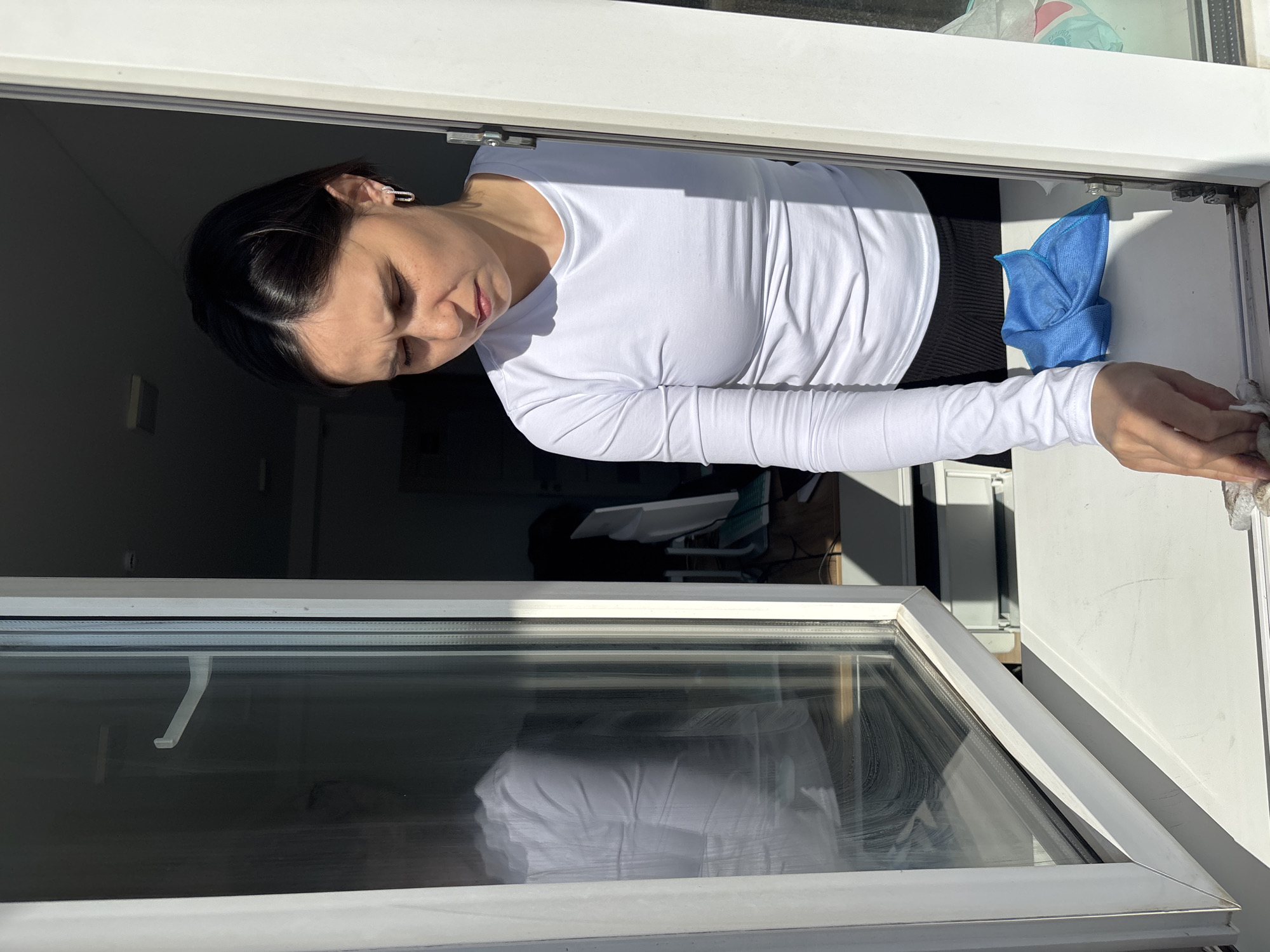 A woman in a white long-sleeved shirt is cleaning a window, holding a cloth in her right hand. A blue microfiber cloth is tucked between the window frame. The room visible through the window appears to contain a desk and computer monitor. The location is not discernible from the image, and there is no indication it is in Izhevsk, Russia.
