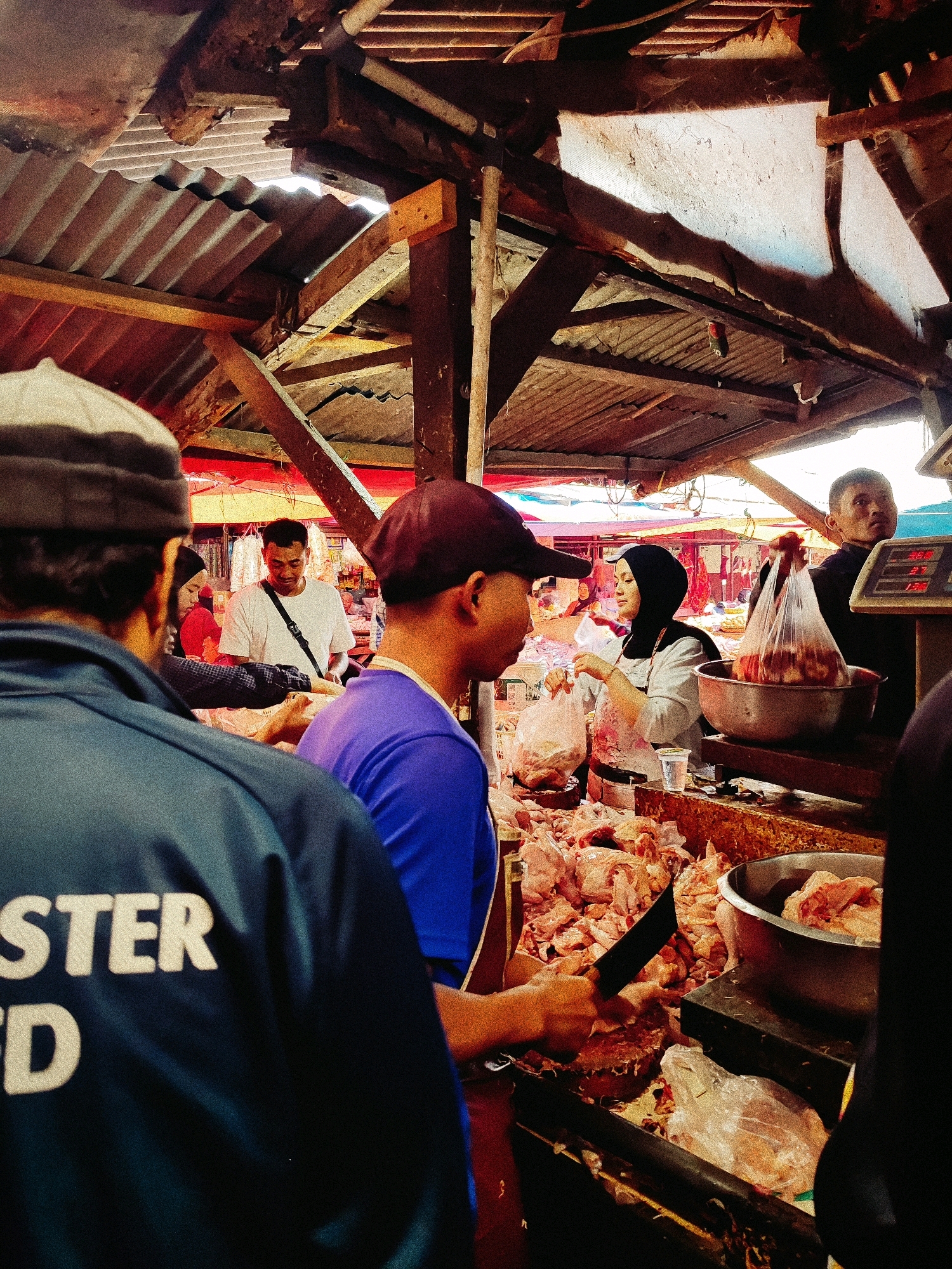 The image depicts a bustling outdoor market scene in Garut, Indonesia, likely a butcher's stall. The foreground is dominated by the backs of two men, one wearing a dark blue jacket with white lettering that appears to say "STER D", and another in a vibrant purple t-shirt. Between them, a butcher in a maroon cap is actively chopping meat with a large knife on a wooden cutting board. He is surrounded by piles of raw meat, predominantly chicken pieces.
Behind the butcher, a woman wearing a black hijab and a white top with red accents is handling plastic bags, possibly packaging meat for customers. She is interacting with a man in a white t-shirt who is looking down at the transaction. To the right of the woman, another man, partially obscured, is standing near a scale. The market stall itself is constructed from rough wooden beams and covered with corrugated metal roofing, suggesting a rustic and traditional setting. The overall atmosphere is one of active commerce, with an emphasis on fresh meat sales. The lighting is somewhat dim, with natural light filtering through gaps in the roofing, creating shadows and highlighting certain areas.