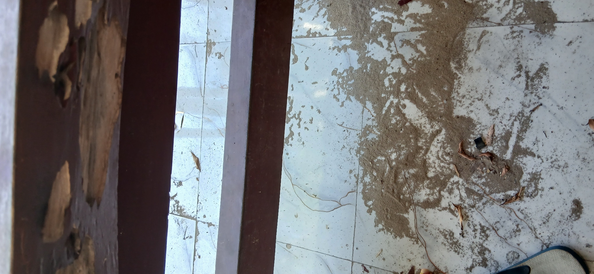 A close-up, downward-facing shot captures a weathered, dark brown wooden beam positioned horizontally in the foreground, with its surface exhibiting signs of peeling paint and exposed wood. Below this beam, a tiled floor extends into the background, appearing to be made of light-colored, possibly marble or ceramic tiles with subtle veining. A significant portion of the floor, particularly in the mid-ground and extending towards the foreground, is covered with a thick layer of sand and dirt, interspersed with dried leaves and debris. A single, brown dried leaf is visible on the tiled surface above the sand deposit. In the lower left corner, a portion of a flip-flop or sandal, predominantly white with a dark blue strap, is visible. The lighting suggests an outdoor or well-lit indoor environment. No individuals are present in the frame. The scene appears to be an outdoor area, possibly a balcony, porch, or patio.