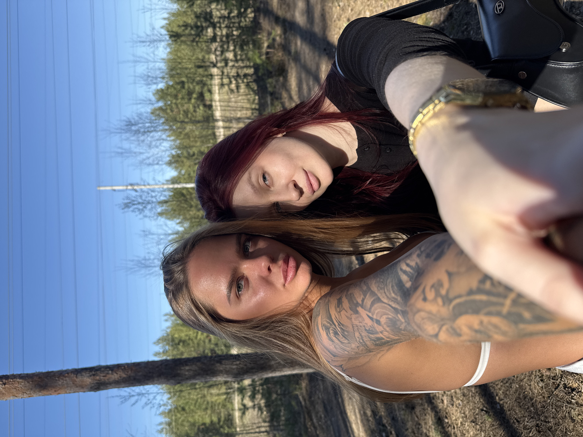 Two women pose for a selfie outdoors. One woman has blonde hair and a tattoo on her left arm and shoulder. She wears a white top. The other woman has red hair and wears a black top. The background includes trees, a clear blue sky with power lines. The location is likely an outdoor area in Nizhny Novgorod, Russia.