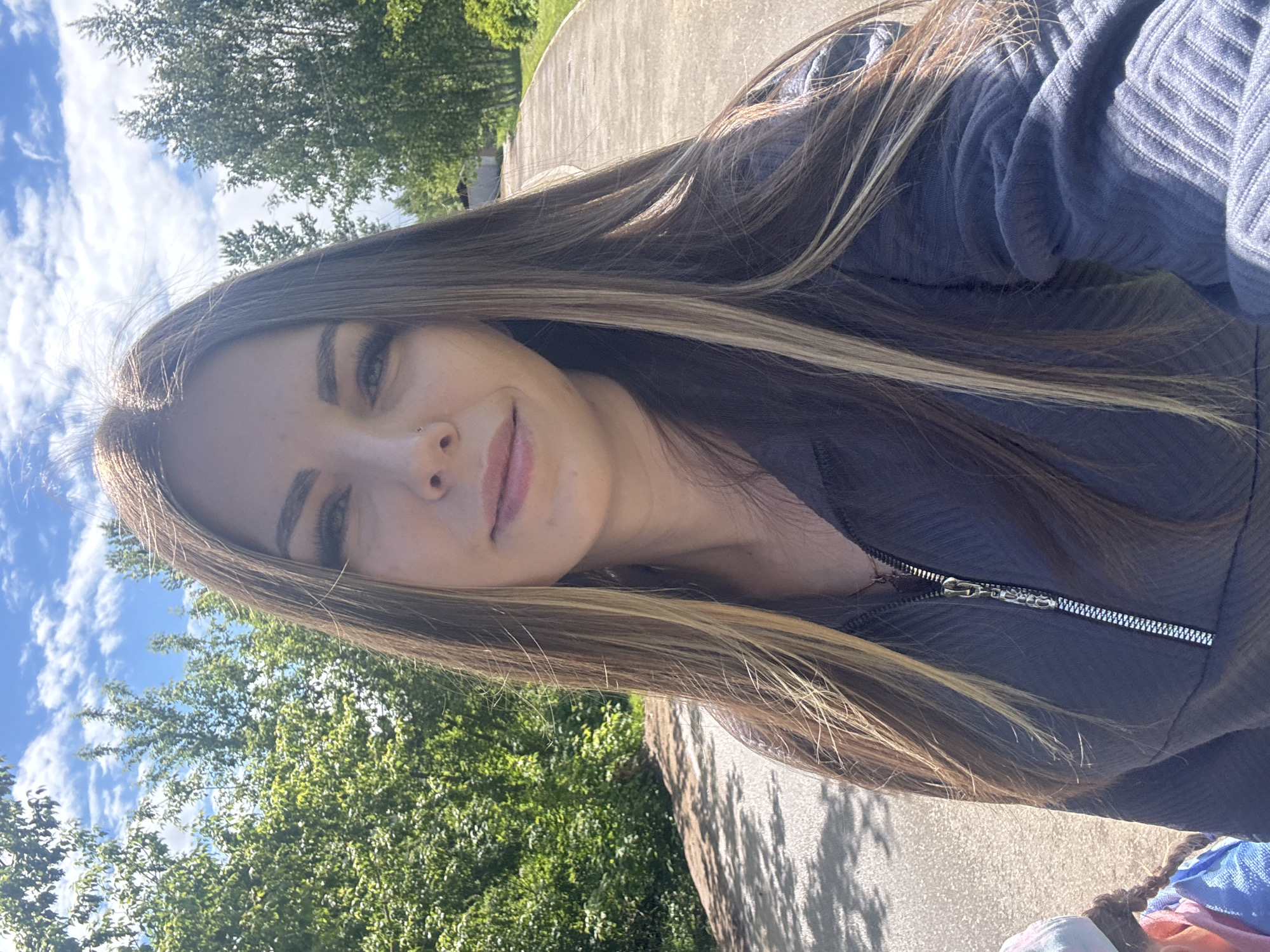 A young woman with long, brown and blonde hair, possibly of Eurasian descent, is taking a selfie outdoors. She is wearing a dark blue, zip-up sweater. A tree and blue sky with clouds are visible in the background. The image was not taken in Shevlyakovo, Russia, as the environment does not match the typical landscape of that location.