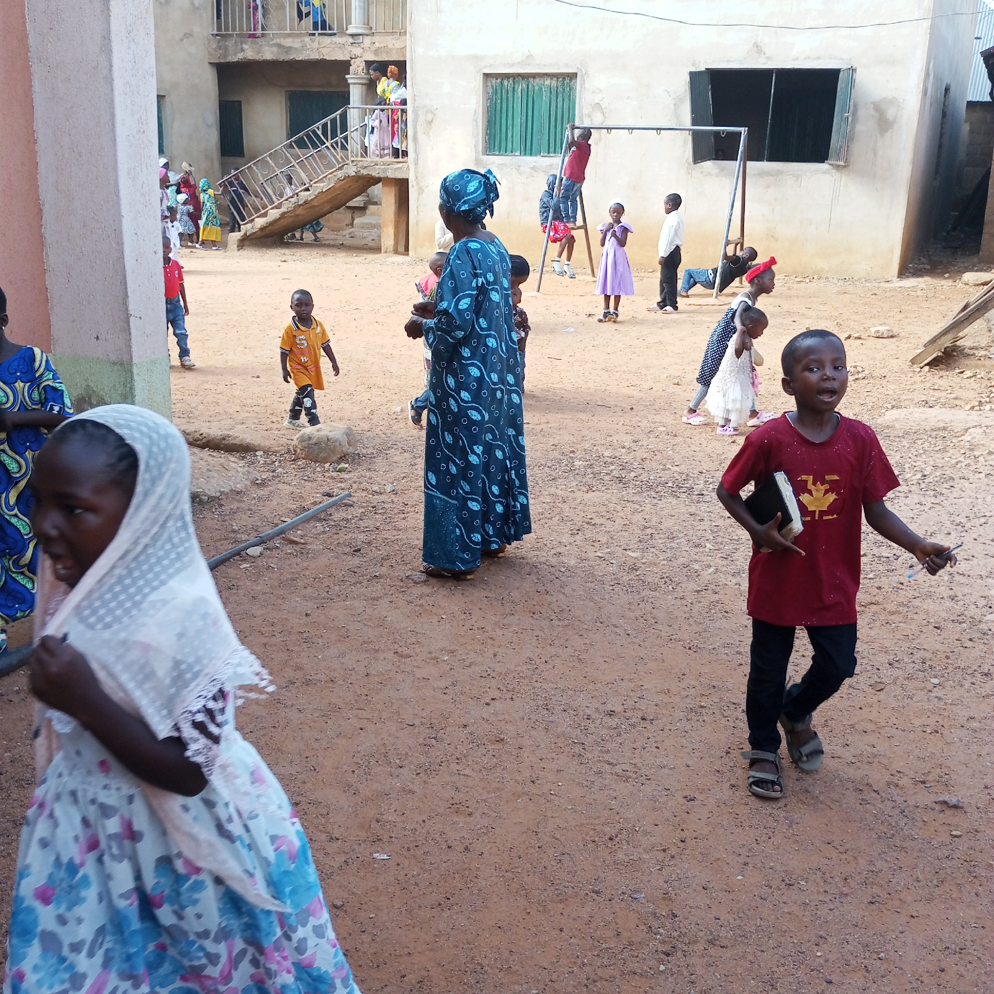 Bukuru, Nigeria: An outdoor scene with a dirt ground surface shows a group of people, primarily children, in a courtyard or open area between buildings. The buildings are of a simple construction, with a concrete structure and windows with bars. Some people are on a balcony or upper level accessed by an exterior metal staircase. A woman in a blue patterned dress and head covering is standing. A child in a white dress with a head covering is in the foreground. A boy in a red shirt is carrying a book and pen. Several other children are playing or standing near a metal play structure, including swings and climbing bars.