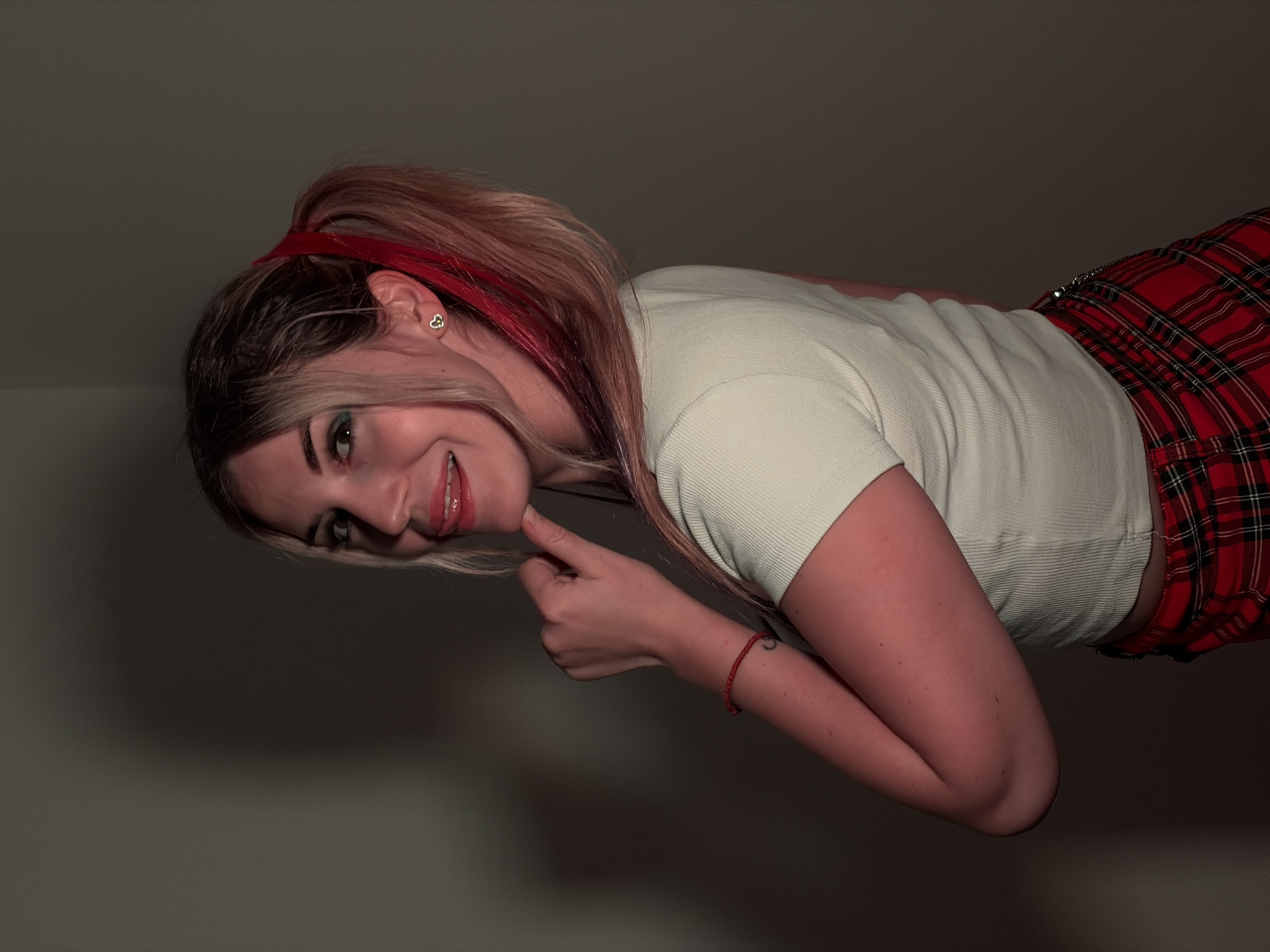 A young woman, with blonde hair streaked with pink and red, wearing a white top and a red plaid skirt, is pictured. She is smiling and playing with her hair, her expression conveying a sense of playfulness. She has a small heart-shaped earring in her left ear and a red bracelet on her right wrist. The background is a plain, dark wall, with no discernible features. The lighting is dim, suggesting an indoor setting, possibly at night. The overall mood is lighthearted and casual. There is no visible text in the image. The context of Budva, Montenegro, is provided but not visually evident in the image itself.