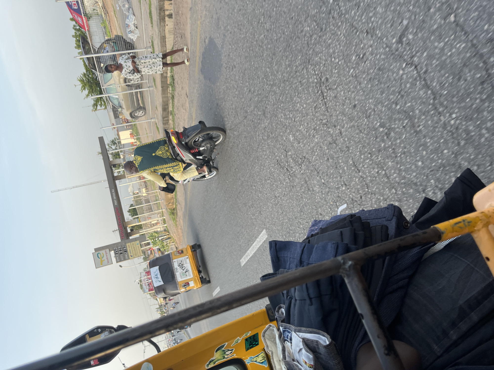 In an outdoor urban scene, a bright yellow auto-rickshaw is seen from the passenger's perspective, with its metallic frame and folded dark blue fabric occupying the foreground. In the mid-ground, a motorcycle rider wearing a green and gold patterned robe and yellow pants is seen from the rear, heading away from the camera. To the right of the motorcycle rider, a young female stands on the side of the road, facing the camera, wearing a white dress with a leaf pattern and white sandals. Behind her, a beige SUV with its trunk open is parked. Further in the background, a gas station with a "TotalEnergies" sign is visible, along with other structures and vehicles. The ground is paved with asphalt, showing some cracks and wear. The sky above is clear and light blue, suggesting it is late afternoon.