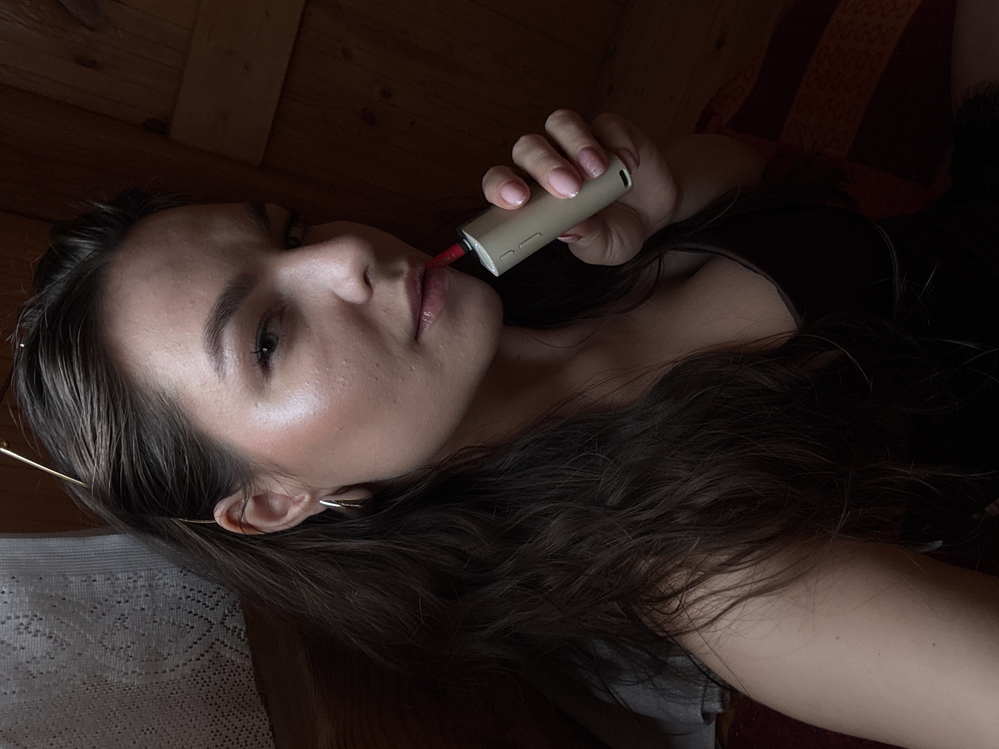 A medium shot captures a young woman with dark brown hair and fair skin indoors. She is wearing a dark camisole and a silver hoop earring in her left ear. Her head is tilted slightly to her right, and she is holding a beige electronic cigarette device to her lips, inhaling. Her right hand is holding the device. The background consists of wooden paneling and a section of lace curtain. The lighting is warm and dim, suggesting an interior setting. The image is a selfie taken from a close-up perspective.