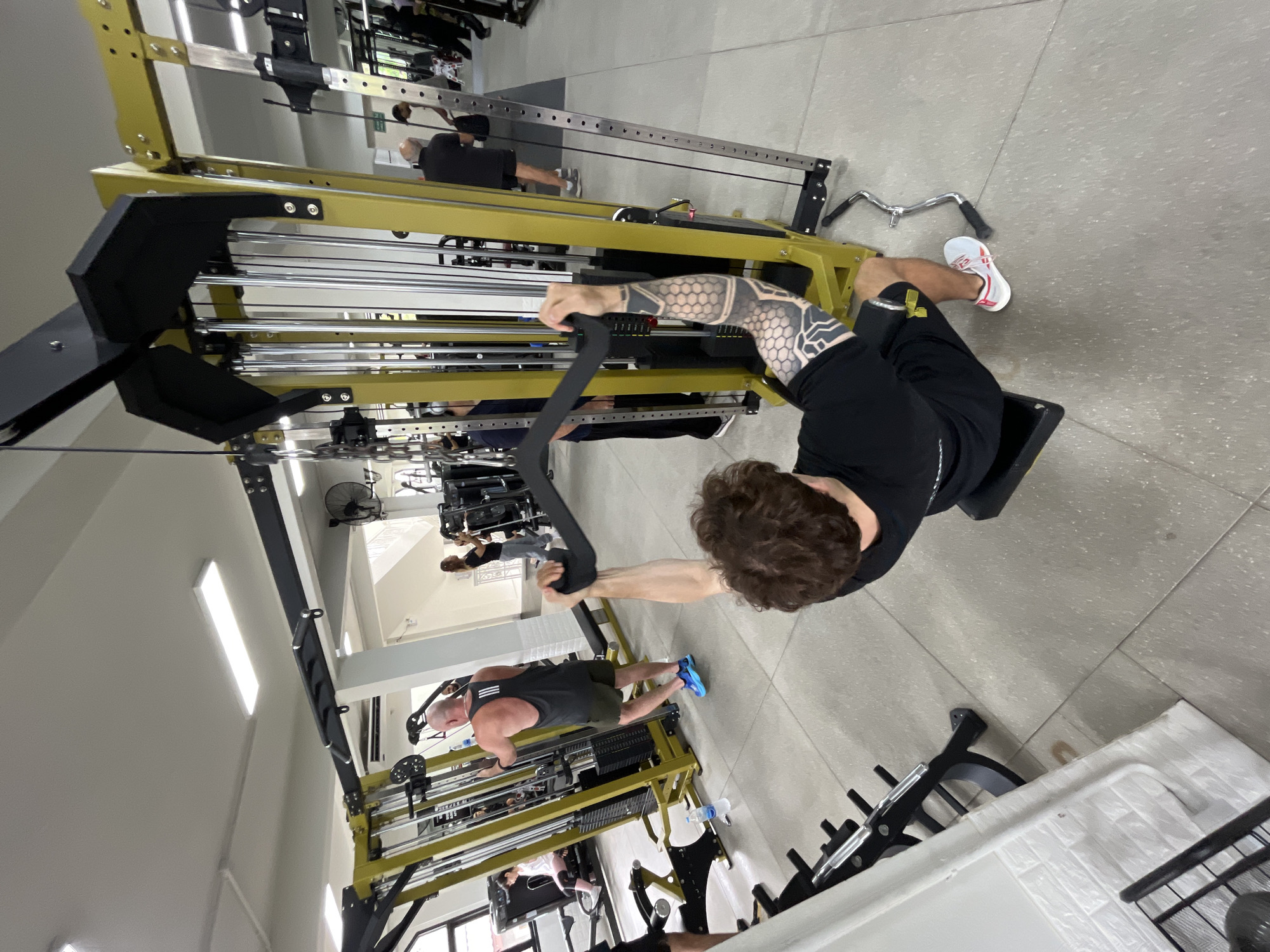 The image depicts the interior of a gym located in Chiang Mai City Municipality, Thailand. In the foreground, a man with curly brown hair and extensive arm tattoos is performing a lat pulldown exercise on a yellow and black weight machine. He is wearing a black t-shirt and black shorts with white and red athletic shoes. To the left and slightly behind him, another man wearing a black tank top and green shorts, with a visible tattoo on his back, is also using a weight machine. In the middle ground, several other individuals are visible engaging in various exercises. One woman in the background is operating a leg press machine. The gym features rows of weight machines and equipment, with ample space between them. The flooring is a light gray speckled tile. The walls are painted white, and the ceiling has fluorescent lighting fixtures. Natural light is entering from windows on the left side of the image.