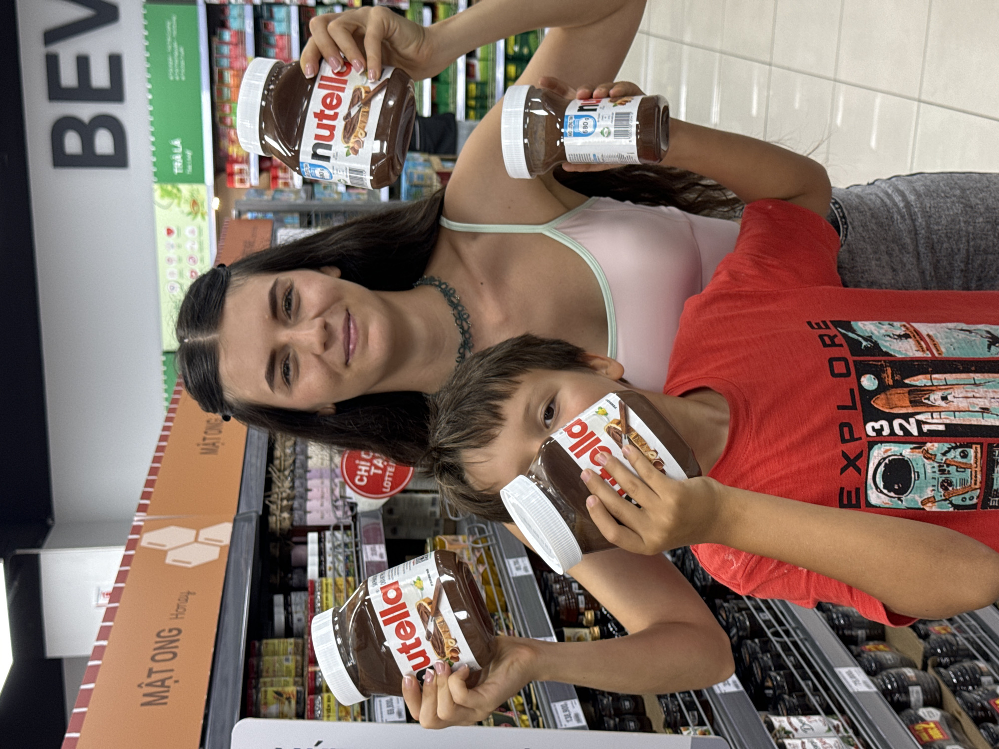 In a shop setting in Phu Thuy Ward, Vietnam, a woman and a child are holding jars of Nutella. The woman is standing and holding a large jar of Nutella in her left hand and a smaller jar in her right hand. She has long dark hair and is wearing a light-colored top with a green trim and a necklace. The child, wearing a red t-shirt, is positioned lower, holding a large jar of Nutella near his face. Shelves stacked with products line the background. Signage is visible, indicating "MẬT ONG" and "TRÀ LA".