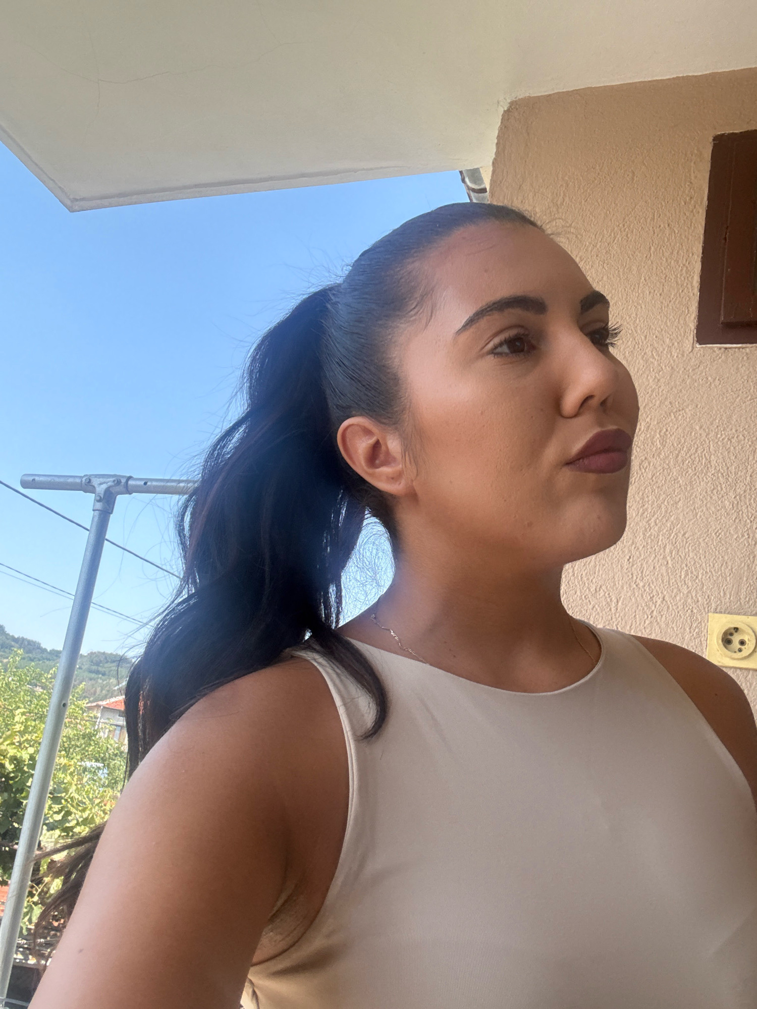 A medium shot shows a woman in profile facing right, her head and shoulders filling the right side of the frame. She has dark hair pulled back into a ponytail, a light-colored tank top, and a thin necklace. Her skin is smooth and her makeup is visible. She is outdoors, with a light blue sky behind her and a beige building to her right, featuring a dark brown window frame and a yellow electrical outlet. A white overhang is visible in the upper left corner. The lower left portion of the image shows a metal drying rack and some green foliage. The scene appears to be on a balcony or porch. Strelcha, Bulgaria is a small town known for its mineral springs and rose production.