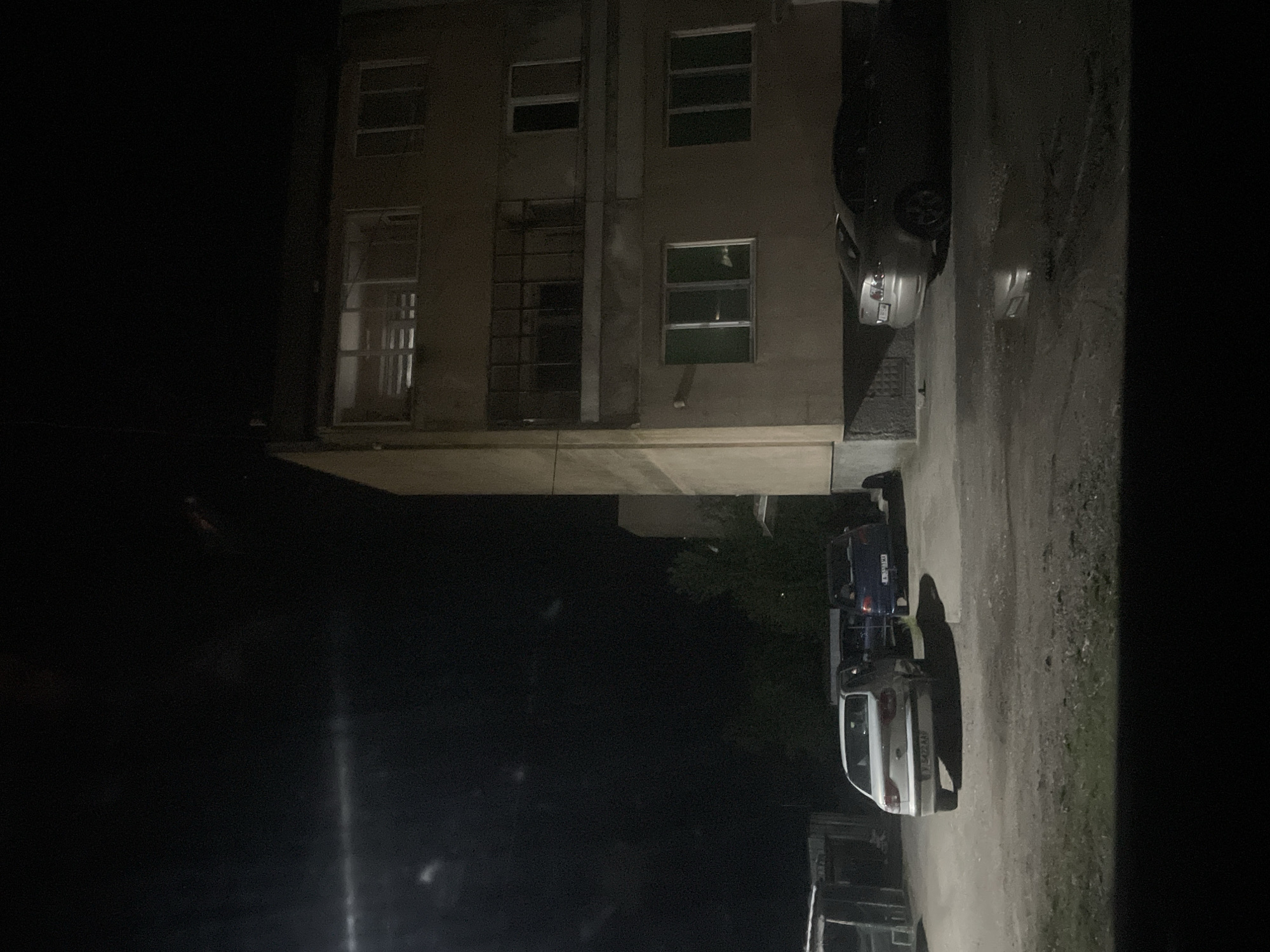 The image depicts a nighttime scene in Debelt, Bulgaria. A three-story building with several windows is visible. Parked on the street alongside the building are a car and a truck, with additional vehicles further down the road. The surrounding environment is dark, with some trees visible in the background.