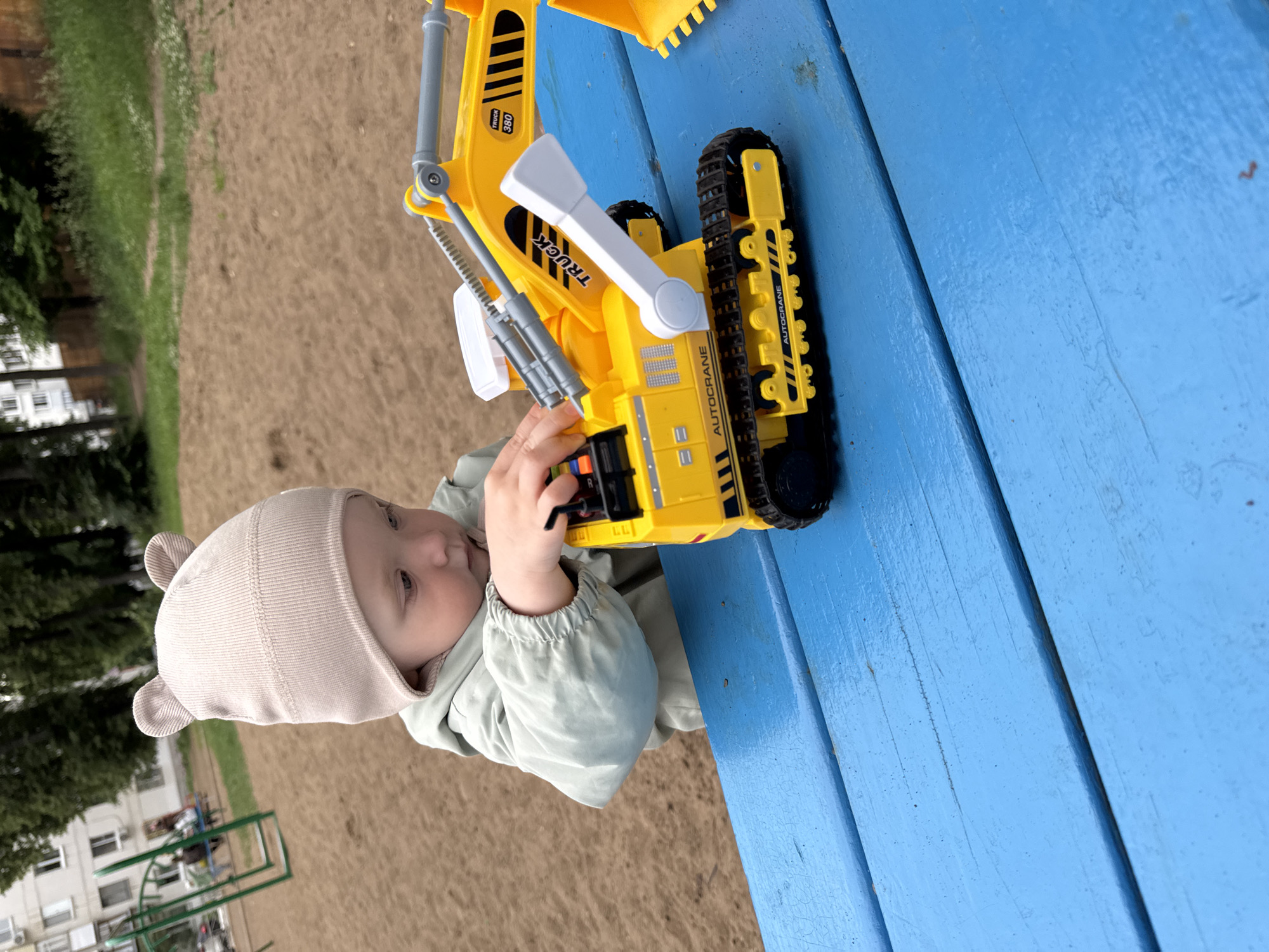 A close-up, eye-level, outdoor medium shot captures a young child playing with a yellow toy excavator on a bright blue wooden picnic table. The child, wearing a light-colored beanie with bear ears and a light green jacket, is focused on manipulating the toy with their right hand. The excavator is detailed, featuring black treads, a movable arm, and text that reads "AUTOCRANE" and "TRUCK 380." The background reveals a sandy playground area with trees and a building suggesting an urban environment, possibly near a residential area in Chaikovsky, Russia. The scene is illuminated by natural daylight, creating a slightly overcast atmosphere.