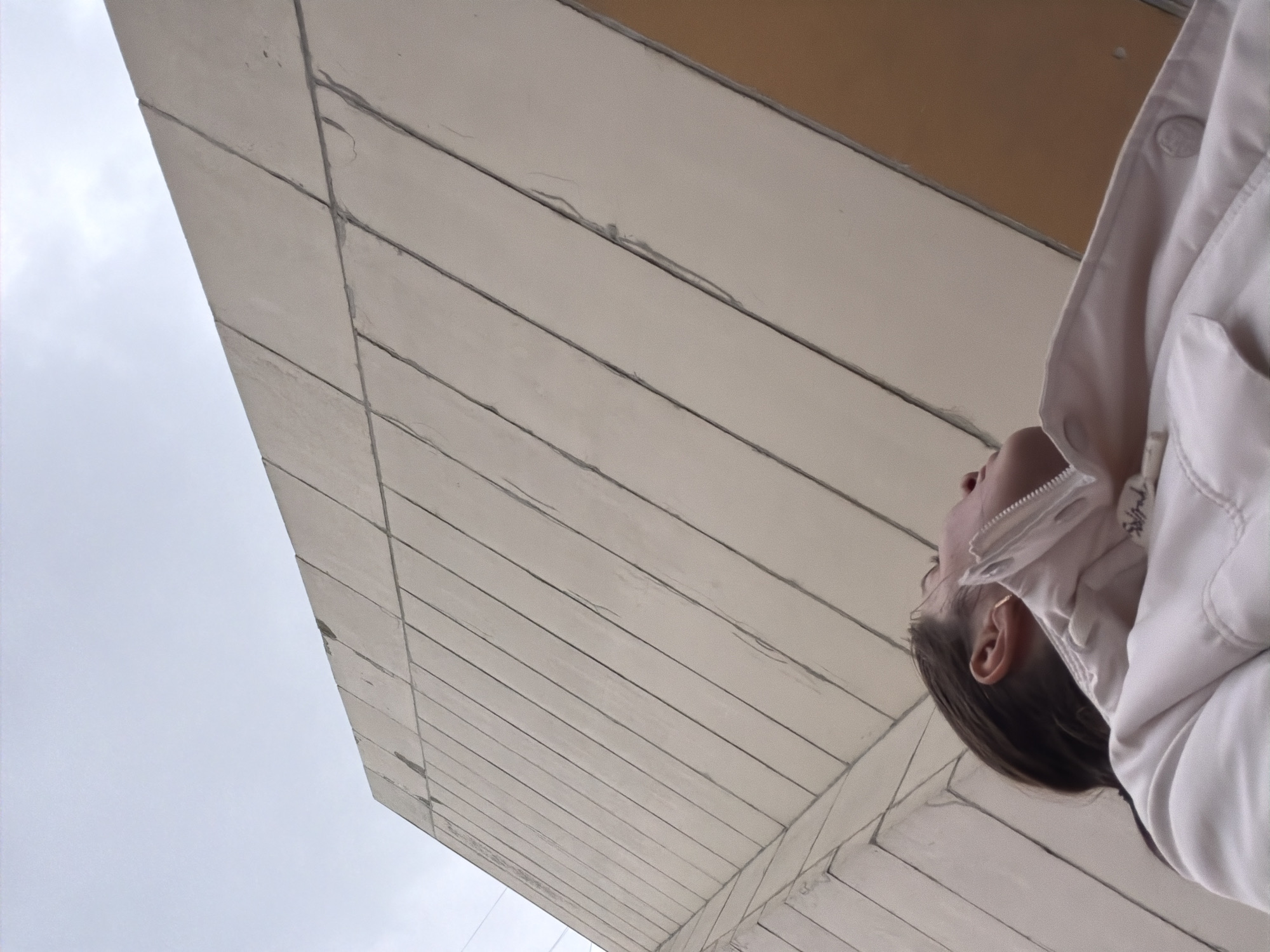 The image shows a person looking up at a building facade in Yanino-1, Russia. The facade is constructed of long, rectangular, light-colored panels with visible seams. The sky is overcast. The person is wearing a light-colored jacket and has dark hair.