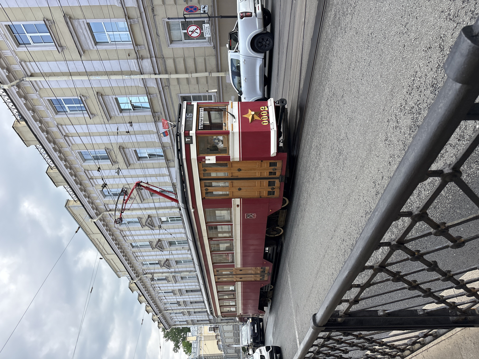 A vintage red and white tramcar with gold accents is stopped on a street in Saint Petersburg, Russia. The tram has the number 0002 displayed prominently with a gold star next to it. The street is lined with buildings featuring classical architecture. Overhead electrical lines power the tram. The sky is overcast. A white pickup truck and other vehicles are parked along the street. A metal fence is visible in the foreground. The tram appears to be a tourist attraction.