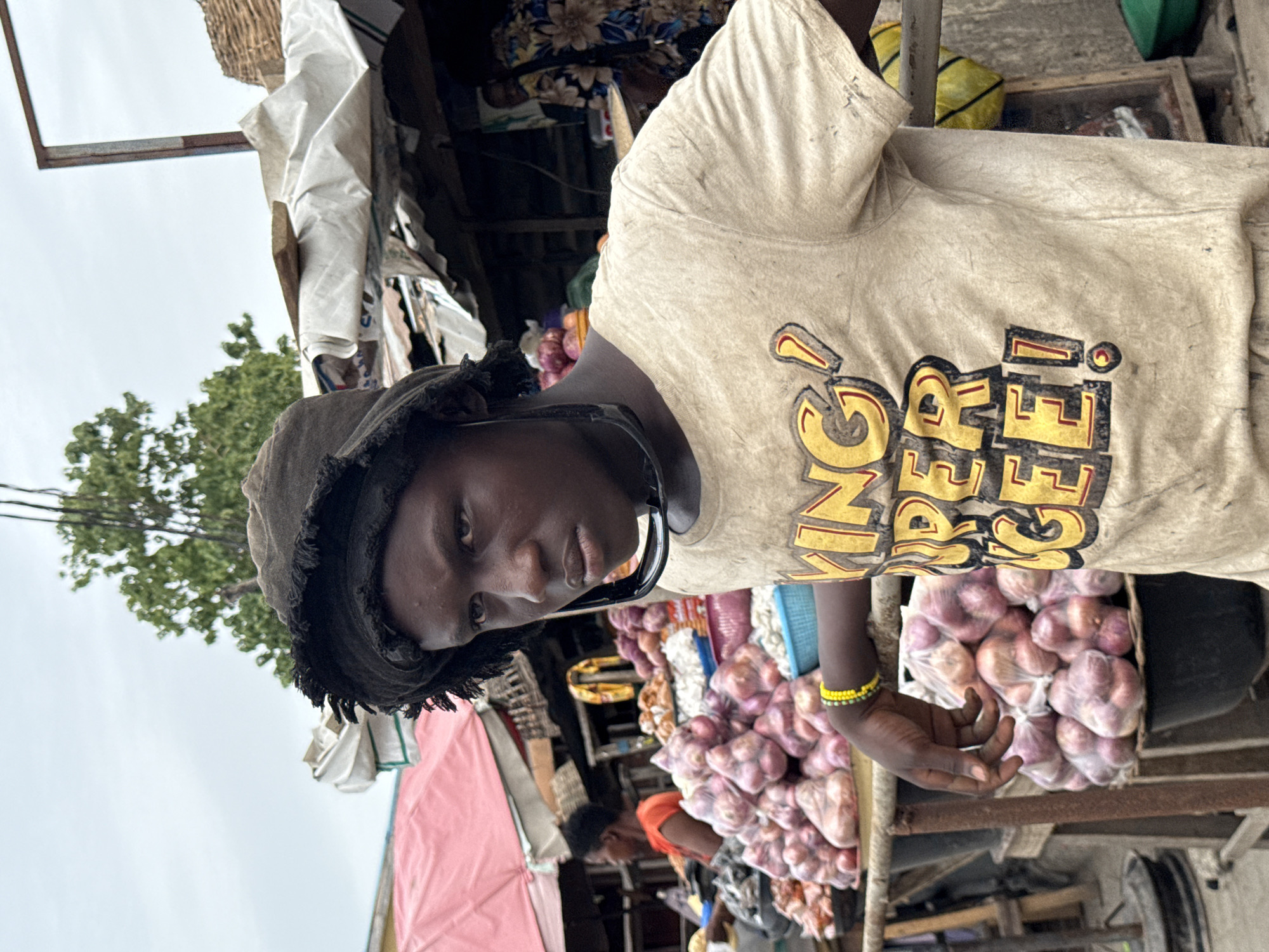 A young person wearing a fringed hat and a t-shirt with the words "KING SELLER!" in yellow and black is standing in a market. They have a yellow bracelet on their right wrist. Stacks of onions wrapped in clear plastic are in front of them. The market appears to be outdoors, and other stalls are visible in the background, some with pink canopies. A tree is visible in the upper left corner. Based on the visual style and details of the stalls, this is likely a market setting in Lagos, Nigeria.
