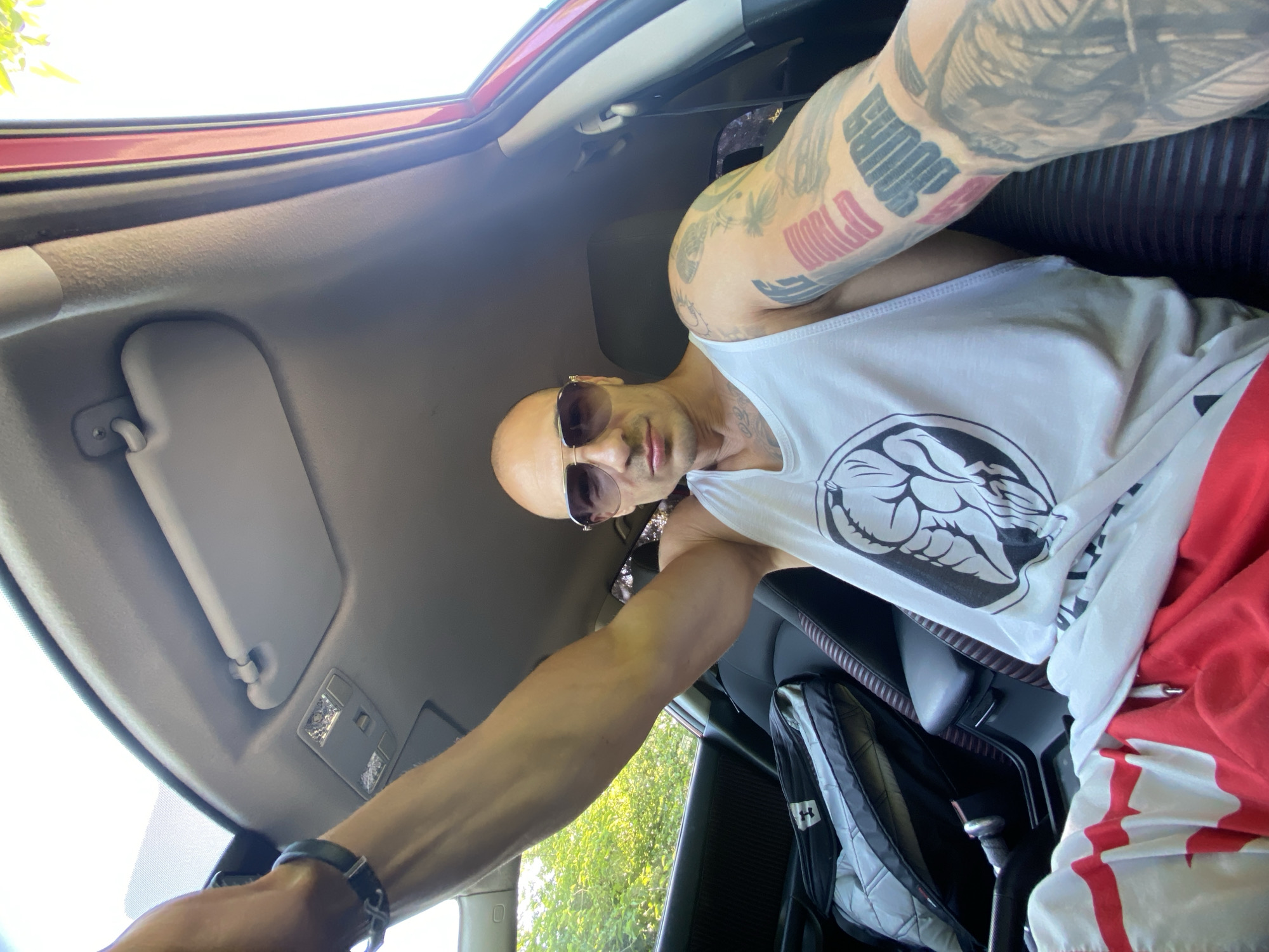 A bald man with sunglasses and tattoos on his arm is sitting in the passenger seat of a red car. He is wearing a white tank top with a graphic design and red and white patterned shorts. A black and grey bag is visible on the back seat. The car appears to be parked outdoors, with trees and foliage visible through the open window. The lighting suggests it is daytime, with the sun casting a bright light. The image was taken from a low angle, looking up at the man and the car's interior.