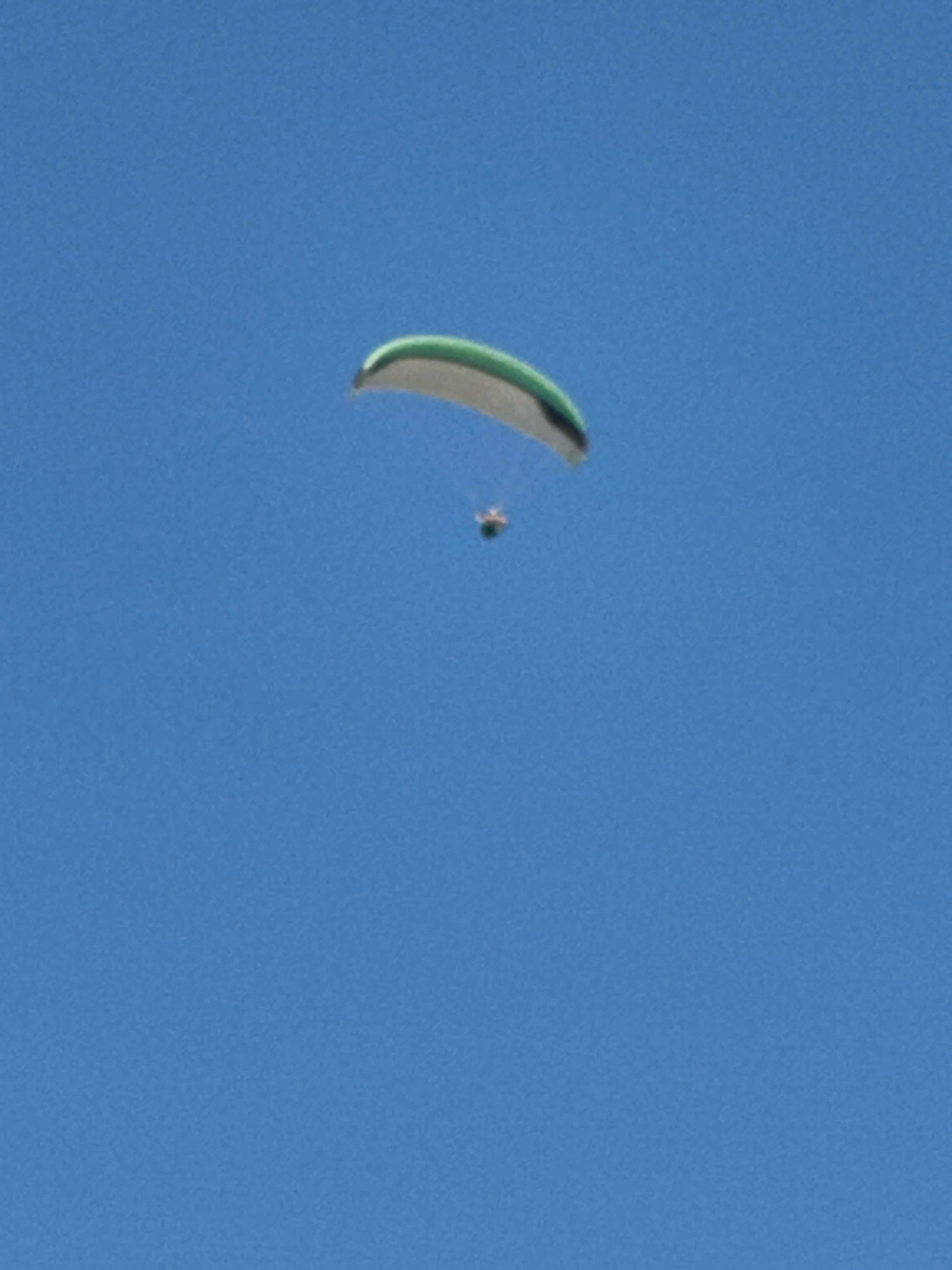 A paraglider is seen in mid-flight against a clear blue sky. The paraglider is green and white and appears to be descending. A person is visible attached to the paraglider. The image does not provide any information about the location in Boreti, Montenegro.
