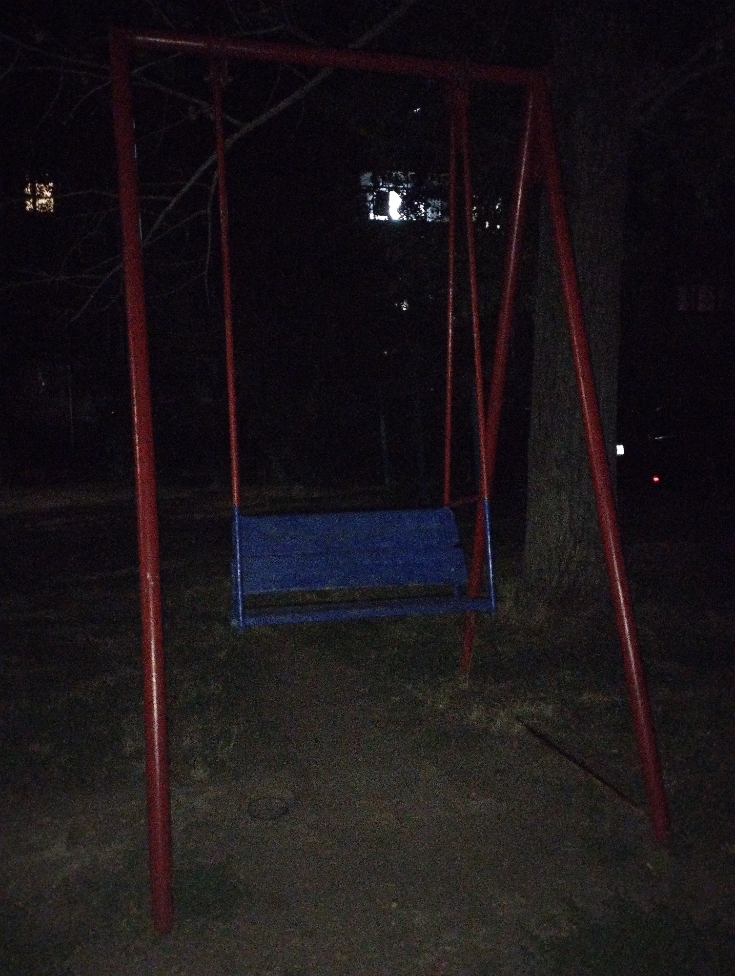 The image depicts a swing set in what appears to be a dimly lit outdoor area, possibly a park or backyard, at night. The swing set has a red metal frame and a blue seat. The background is dark, with faint lights visible in the distance, suggesting it is in or near a populated area. There are no people or animals present in the image, and no obvious actions are taking place. The ground around the swing set is covered with dirt and grass. The lighting conditions are poor, with significant noise in the image, making fine details difficult to discern. The location in Belaya Kalitva, Russia, is not visually indicated within the image itself.