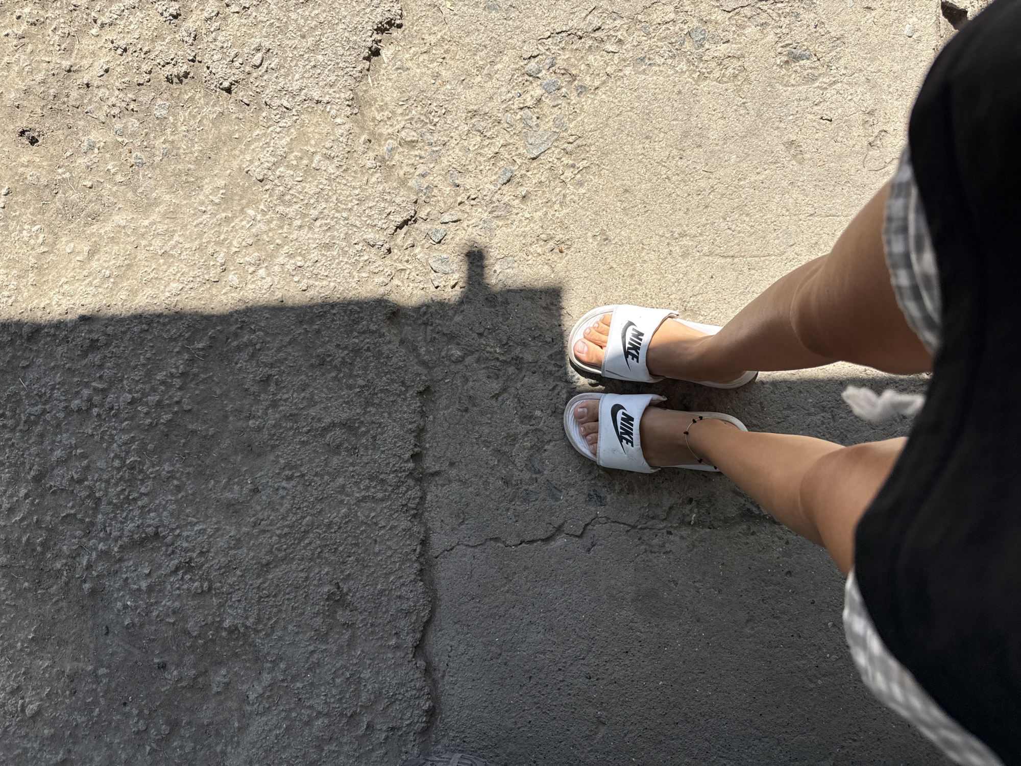 In Basarabeasca, Moldova, a person stands on a cracked, sun-drenched asphalt surface. The perspective is from above, focusing on the lower legs and feet. The individual is wearing white Nike slides and a black skirt with a checkered waistband. A dark shadow stretches diagonally across the ground from the left side of the frame, obscuring a portion of the asphalt. The ground is rough and uneven, with visible debris and cracks. The lighting suggests it is daytime, with harsh sunlight casting sharp shadows.