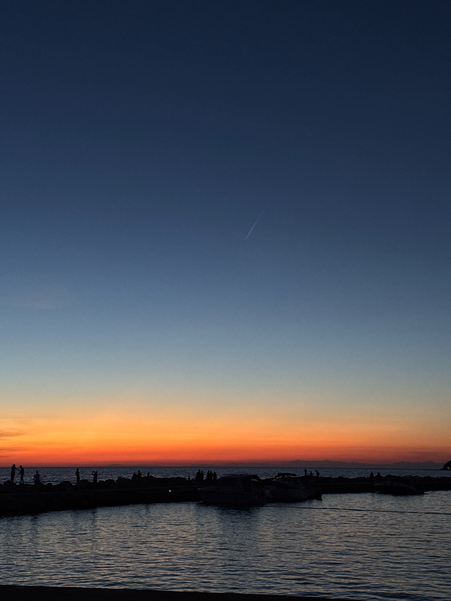 This is a vertical, full-frame, outdoor, daytime photograph captured during sunset. The scene depicts a body of water with a dark, rocky breakwater in the foreground, where numerous people are silhouetted. Several boats are docked within the breakwater. The sky transitions from a deep blue at the top to vibrant orange and yellow hues closer to the horizon. A thin white contrail from an aircraft is visible in the upper portion of the sky. The location is identified as Novigrad, Croatia.