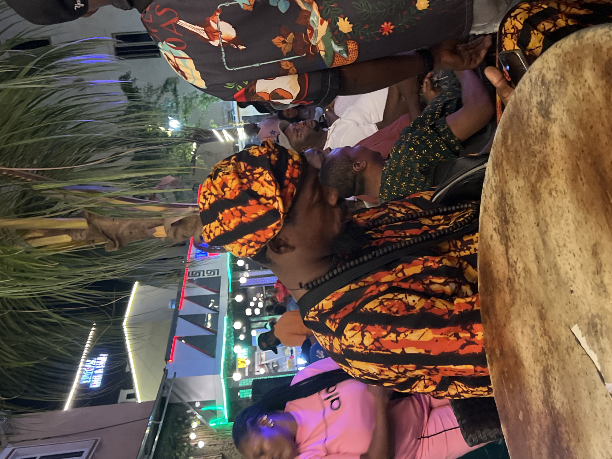The image depicts an outdoor nighttime scene in the city of Oba, Nigeria. A group of people are gathered, some appearing to be engaged in a musical performance. In the foreground, a man wearing a colorful, patterned head wrap and a striped shirt is seated, his head bowed, seemingly playing a drum that occupies the bottom portion of the frame. To his left, a woman in a pink t-shirt stands, her arm visible. In the background, other individuals are present, some seated and others standing, amidst a setting illuminated by string lights and neon signage on buildings. The environment suggests a lively social gathering or entertainment venue.