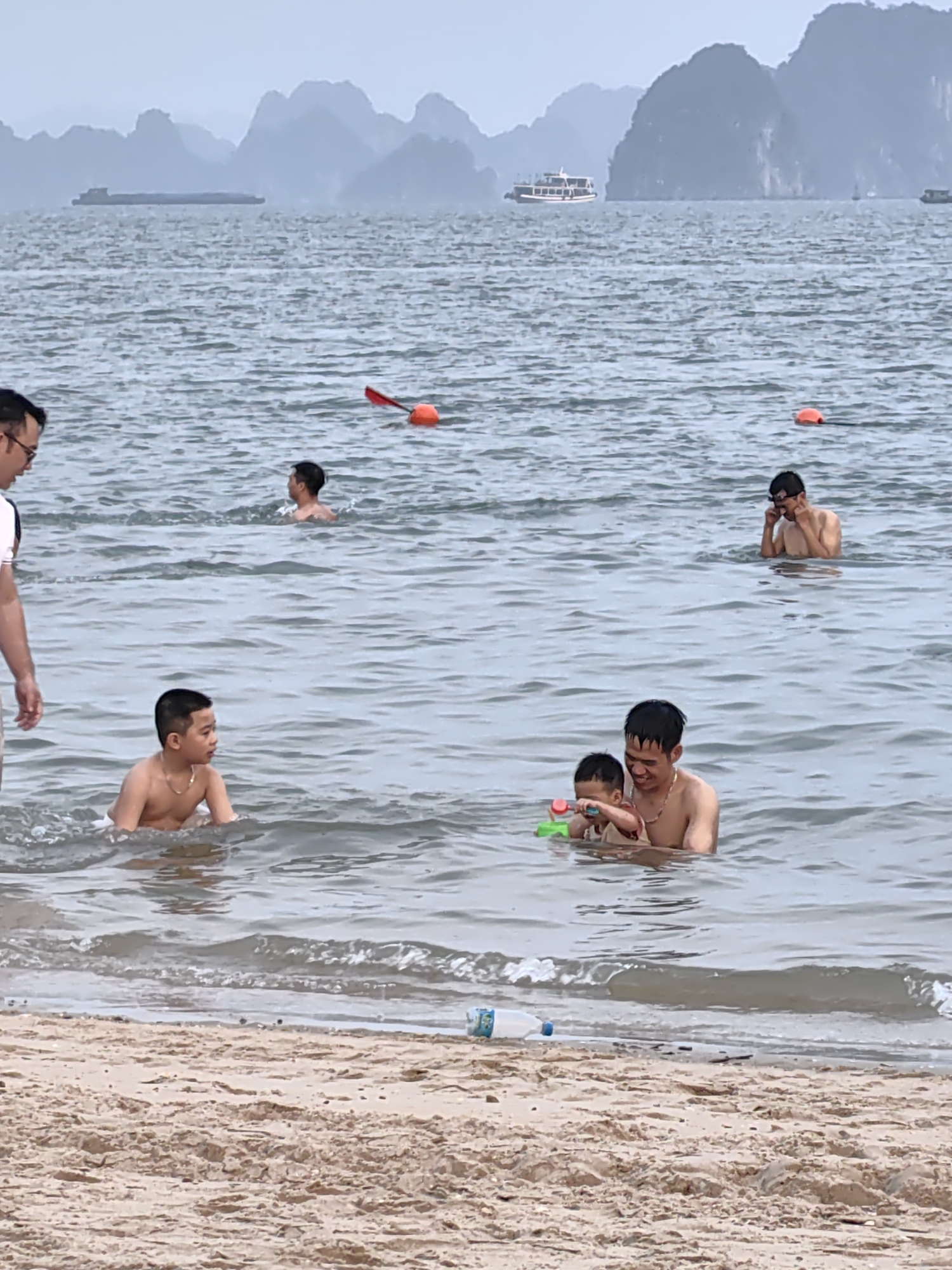 The image depicts a scene at a beach in Bai Chay Ward, Vietnam. Several people are in the water. In the foreground, a boy is wading in the water. Nearby, an adult male is holding a toddler, and both are playing with a green and red toy. Further back, two more individuals are visible in the water, and another adult is partially visible on the edge of the beach. Several orange buoys are floating in the water. In the background, there are distant islands and two boats. There is a discarded plastic water bottle on the beach. The overall environment appears overcast and humid.