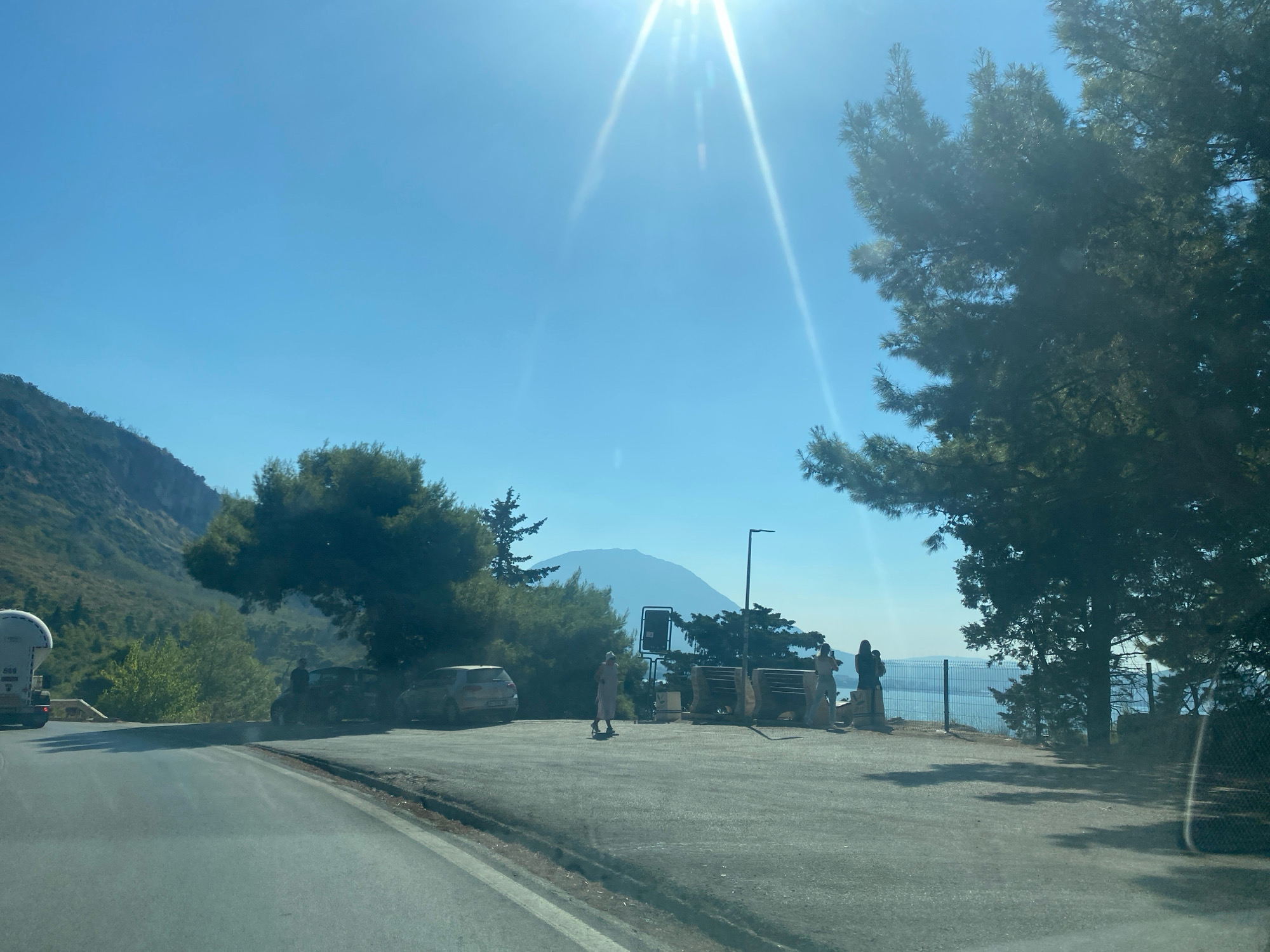 The image depicts a scenic overlook on a sunny day, likely a viewpoint above the town of Sutomore, Montenegro. The foreground features a paved road curving to the left, with a light-colored vehicle parked on the right side of the road. Further back, a parking area is visible with several people standing. One person is walking in the middle of the parking area, dressed in a light-colored outfit. To the right, two individuals are standing near a fence overlooking a body of water, possibly the Adriatic Sea. A large mountain is visible in the distance, shrouded in a light haze. Dense green trees are scattered throughout the scene, framing the vista. The sky is a clear, bright blue with the sun prominently shining, creating lens flare.