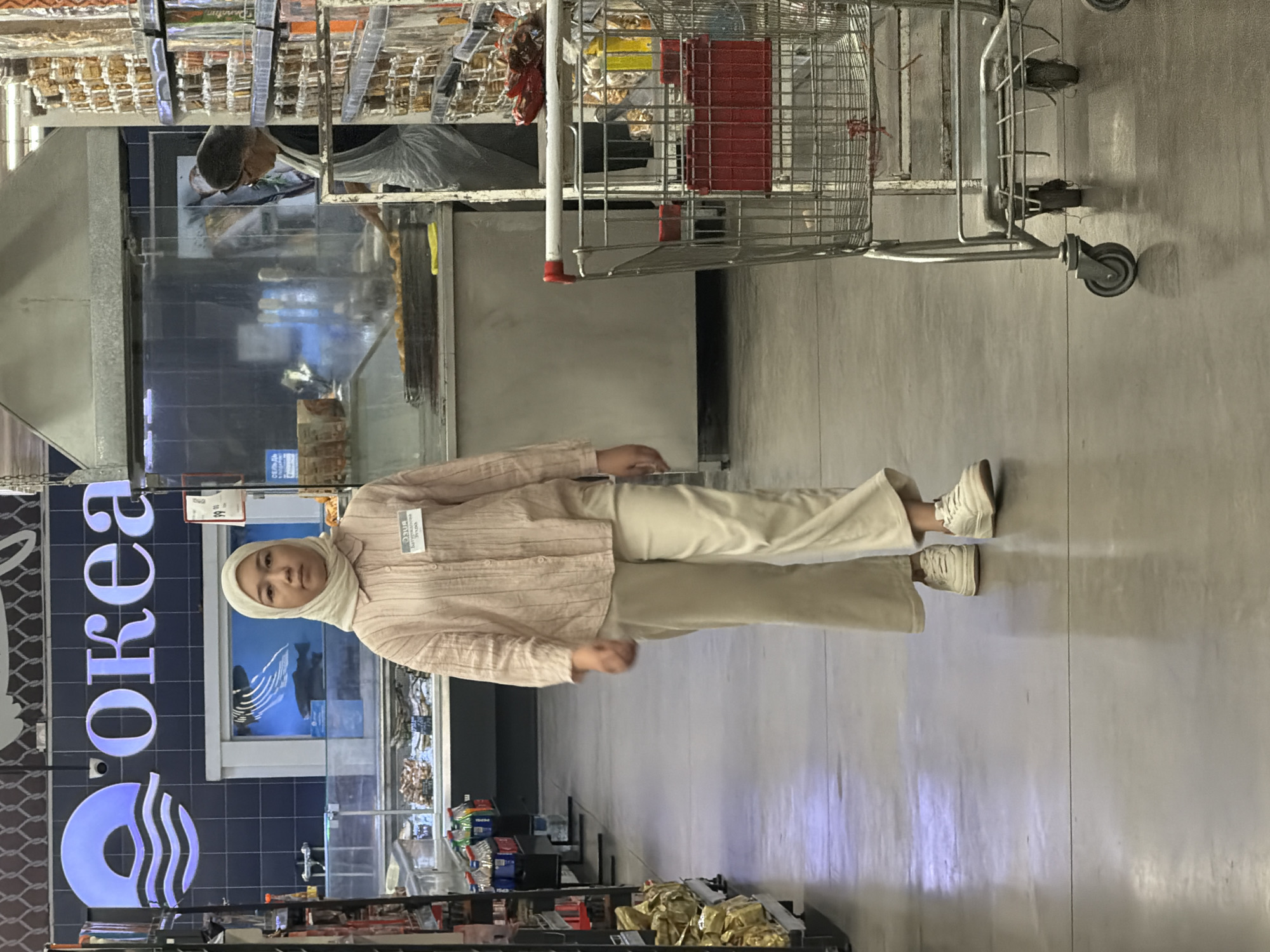 In an indoor supermarket aisle, a young woman wearing a light pink collared shirt, loose cream pants, and white sneakers walks towards the viewer. She also wears a light cream-colored hijab and has a name tag on her shirt. She is looking straight ahead. Behind her, a supermarket aisle displays various food items on shelves. To the right, a metal shopping cart with a red crate is positioned. Further back, behind a glass partition, a person in a hair net and apron is seen working at a food counter. The supermarket aisle is well-lit with fluorescent lighting.