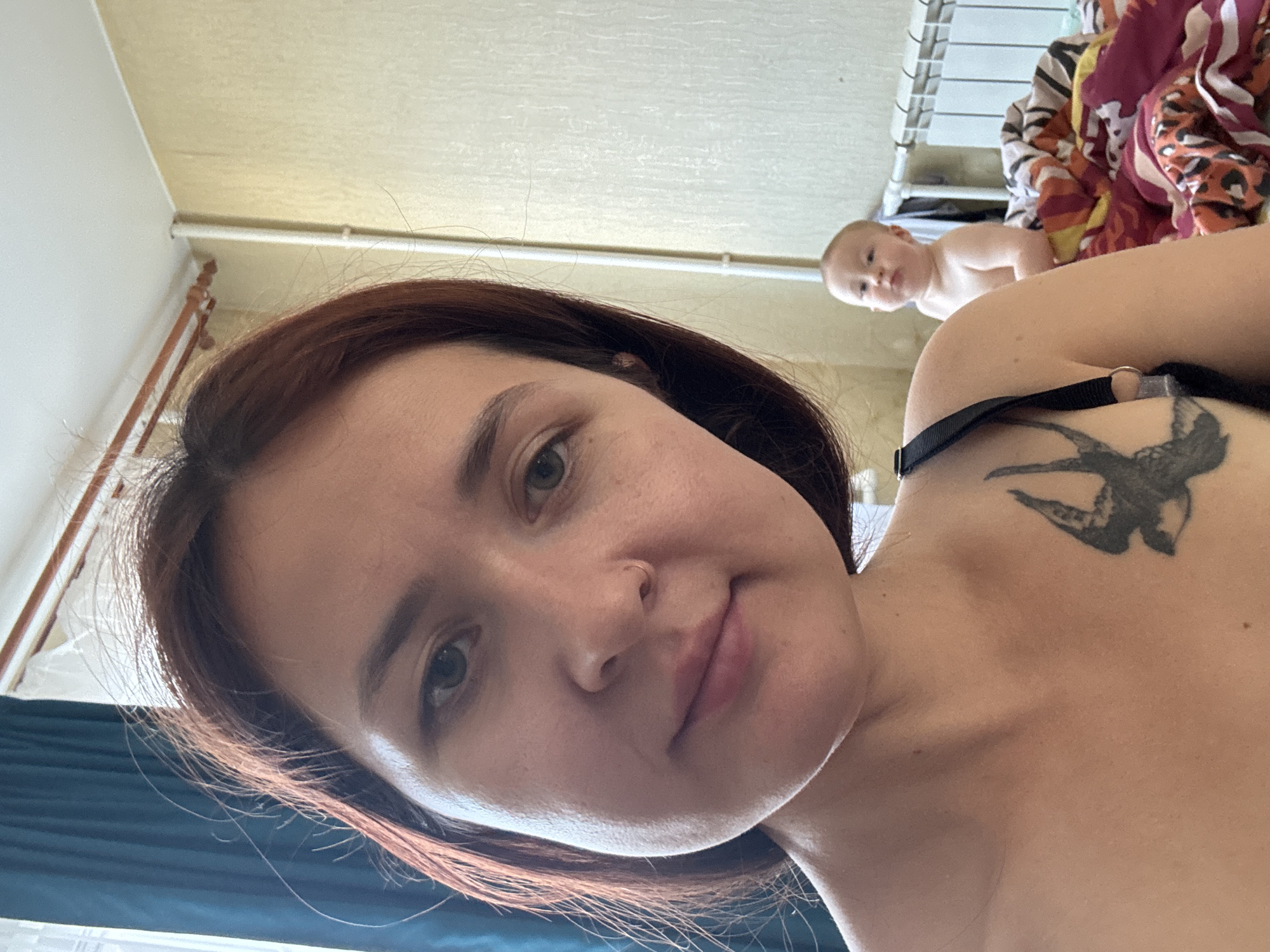 A woman with auburn hair and a tattoo on her left shoulder is taking a selfie indoors. She is in the foreground, with her face angled towards the camera. In the background, a baby is sitting on a bed. The room appears to be a bedroom, with a radiator and curtains visible. The overall lighting suggests it is daytime. The location is identified as Naberezhnye Chelny, Russia.