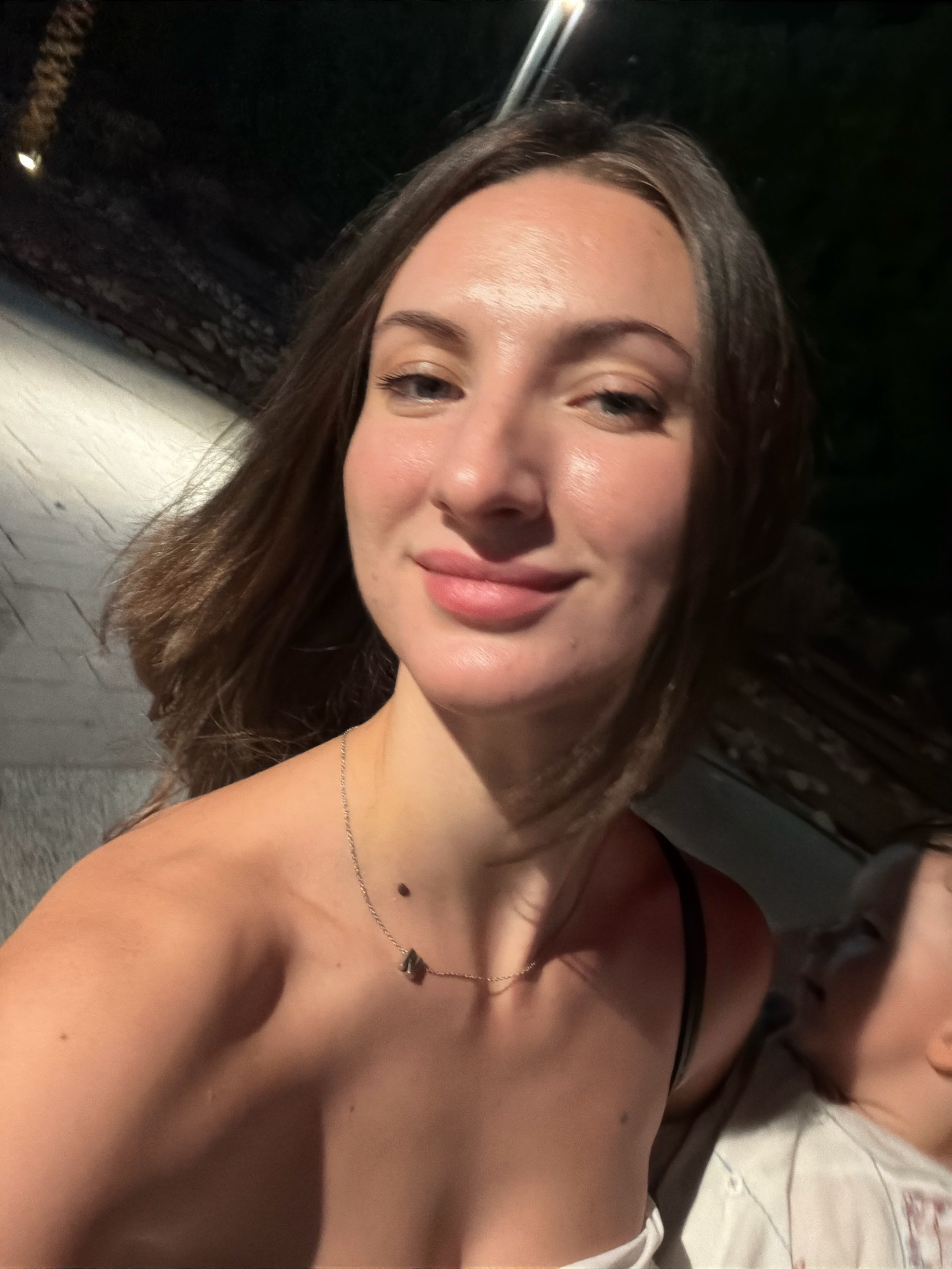 The image shows a woman in the foreground, holding what appears to be a child. The woman is looking at the camera and smiling. She has long brown hair and is wearing a necklace. The child is looking away from the camera. The background is dark and appears to be outdoors. The image is a selfie, taken by the woman. There are no recognizable landmarks or specific details that would indicate the location as Sharm El Sheikh, Egypt. The lighting suggests it is nighttime.