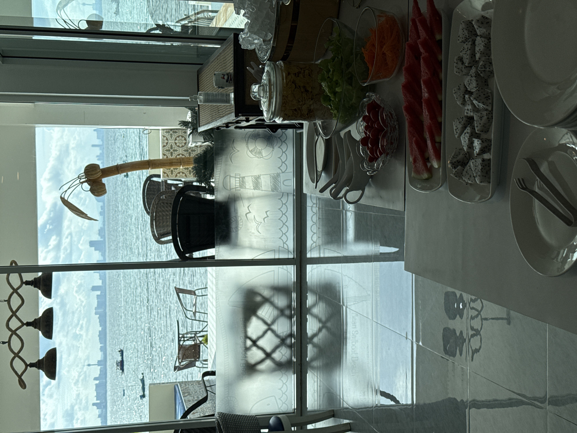 A breakfast buffet is set up indoors with a view of the sea. The buffet includes a variety of fresh fruits like watermelon and dragon fruit, along with salad ingredients such as lettuce, tomatoes, and shredded carrots. There are also condiments and plates available. The view from the window overlooks the sea, with visible land in the distance, and a few small boats. Outside seating is visible. The window pane features images with the words "Dean Sailboat Beach." The city of Pattaya City, Thailand is visible in the background.