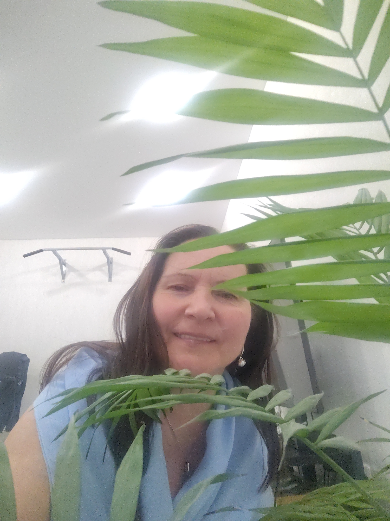 A woman with long brown hair and a blue shirt is partially visible behind a leafy green plant. She is smiling. In the background, there is a white wall with a gray pull-up bar mounted on it. A black object is visible to the left of the woman. The setting is likely indoors, with a bright light overhead. There is no explicit evidence to confirm the location as Sarapul, Russia.