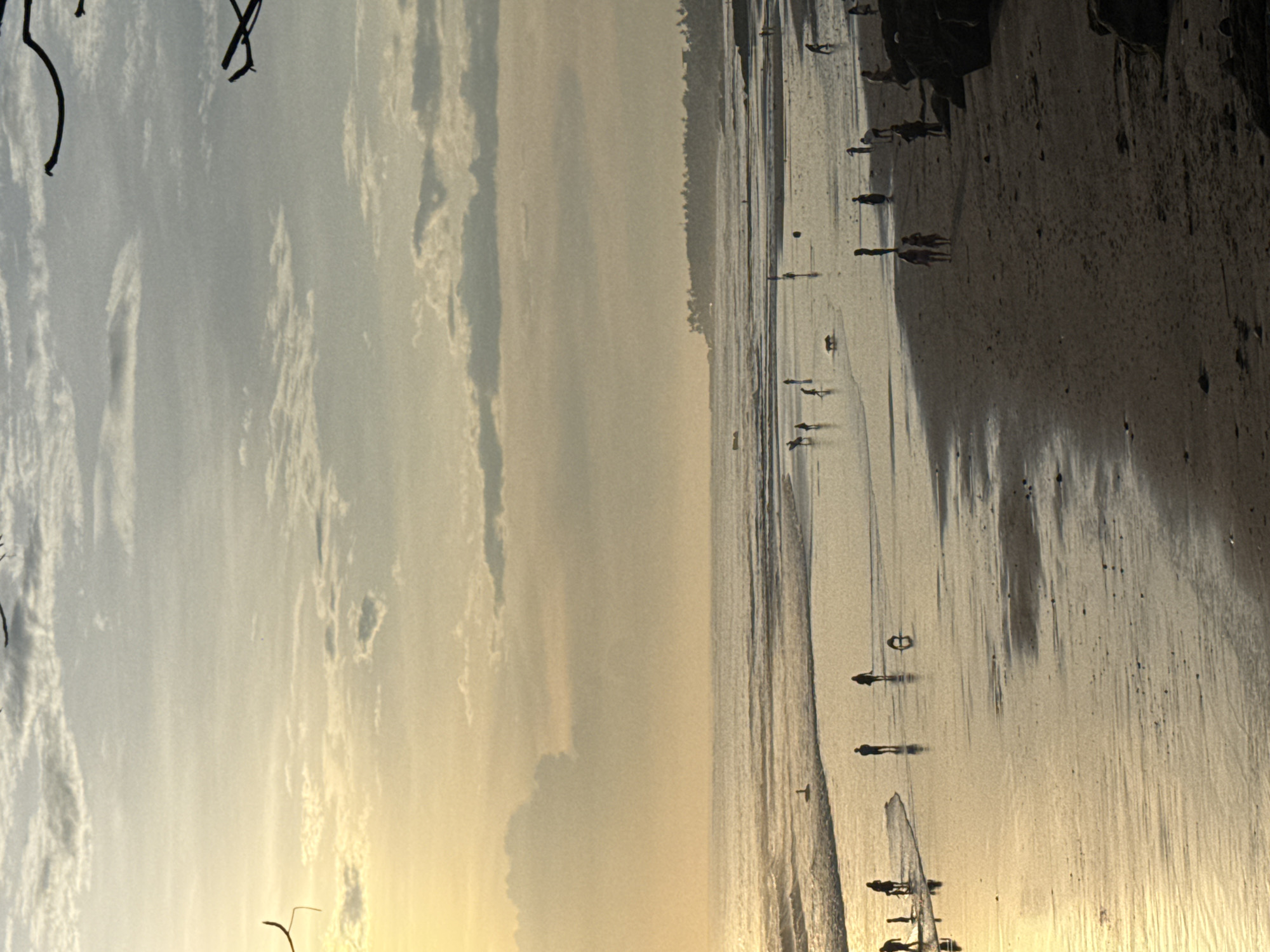 This image captures a serene beach scene at either sunrise or sunset, casting a warm, golden glow across the landscape. The sky, occupying the upper left, displays scattered white clouds against a soft, gradient background that shifts from pale blue to a gentle amber near the horizon.
The wide, flat beach dominates the midground and foreground, meeting a calm body of water, likely the ocean, in the distance. The wet sand at the water's edge acts as a reflective surface, mirroring the sky's light and creating shimmering vertical streaks. Numerous people are depicted as dark silhouettes, scattered across the expansive beach. Most are walking or standing near the waterline, their forms casting long shadows towards the viewer. Some individuals appear further back on the drier sand.
To the right side of the image, a darker, possibly rocky or vegetated area, also in silhouette, defines the edge of the beach. In the top left corner, out-of-focus dark shapes, possibly tree branches or other elements, frame the scene.
The notable details include the soft, backlighting which creates strong contrasts and a tranquil ambiance. There is no visible text. The overall scene conveys a sense of peace and leisure, characteristic of a coastal setting like North Kuta, Indonesia.
