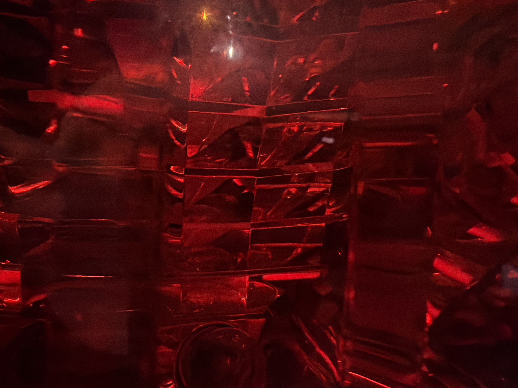 The image shows a close-up, abstract view of a red taillight from a vehicle, likely a car or motorcycle. The red plastic has a textured, faceted surface that refracts and reflects light, creating a pattern of bright red streaks and darker shadowed areas. There are subtle hints of other colors within the reflections, suggesting ambient light from the surroundings. The overall impression is one of intense red illumination and geometric patterns formed by the internal structure of the taillight lens. There are no people, discernible buildings, or specific landmarks visible that would indicate a location in Nha Trang Ward, Vietnam. The image is abstract and focuses solely on the details of the illuminated taillight.