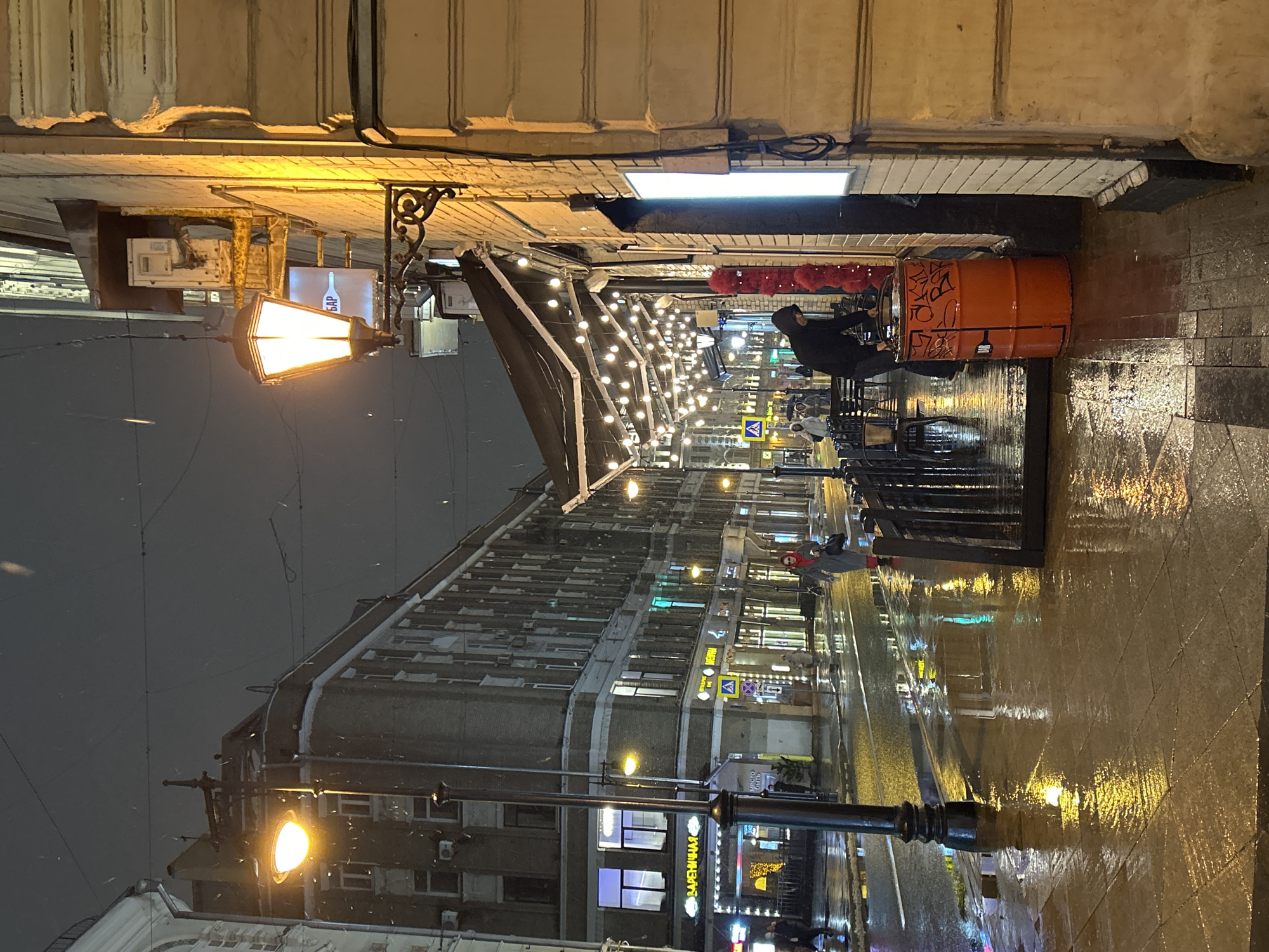 An evening street scene, likely in a city, depicts a wet and reflective pavement under artificial lights, suggesting recent or ongoing rain/light snow. The perspective is looking down a street lined with buildings.
On the right side of the street, an outdoor cafe or bar area is visible, brightly illuminated by overhead string lights. A person in a dark hooded jacket sits at a table, facing away from the viewer. An orange industrial barrel, covered in some graffiti-like Cyrillic text (possibly "ОКТЯБРЬ"), serves as a table or decor. Above the seating area, red puffy decorations are strung. A wall-mounted sign displays a bottle icon and the Cyrillic word "БАР" (BAR). The wet ground in this area reflects the warm glow of the lights.
On the left, a multi-story building with numerous windows extends into the distance. A classic-style street lamp, casting a bright yellow light, stands prominently in the foreground. Another street lamp is visible further down the street. Small white specks are scattered in the air, indicating falling precipitation.
Further down the street, various storefronts are lit, and a yellow diamond-shaped road sign, indicating a pedestrian crossing, is visible. Another sign clearly reads "ВАРЕНИЧНАЯ" (Varenychna), suggesting a dumpling restaurant. The wet street surface dramatically reflects the light from the building signs and streetlamps, creating elongated, shimmering patterns. A few other blurred figures can be seen walking in the distance. The overall atmosphere is urban and somewhat moody, characterized by the contrast between the warm artificial lights and the dark, wet surroundings.