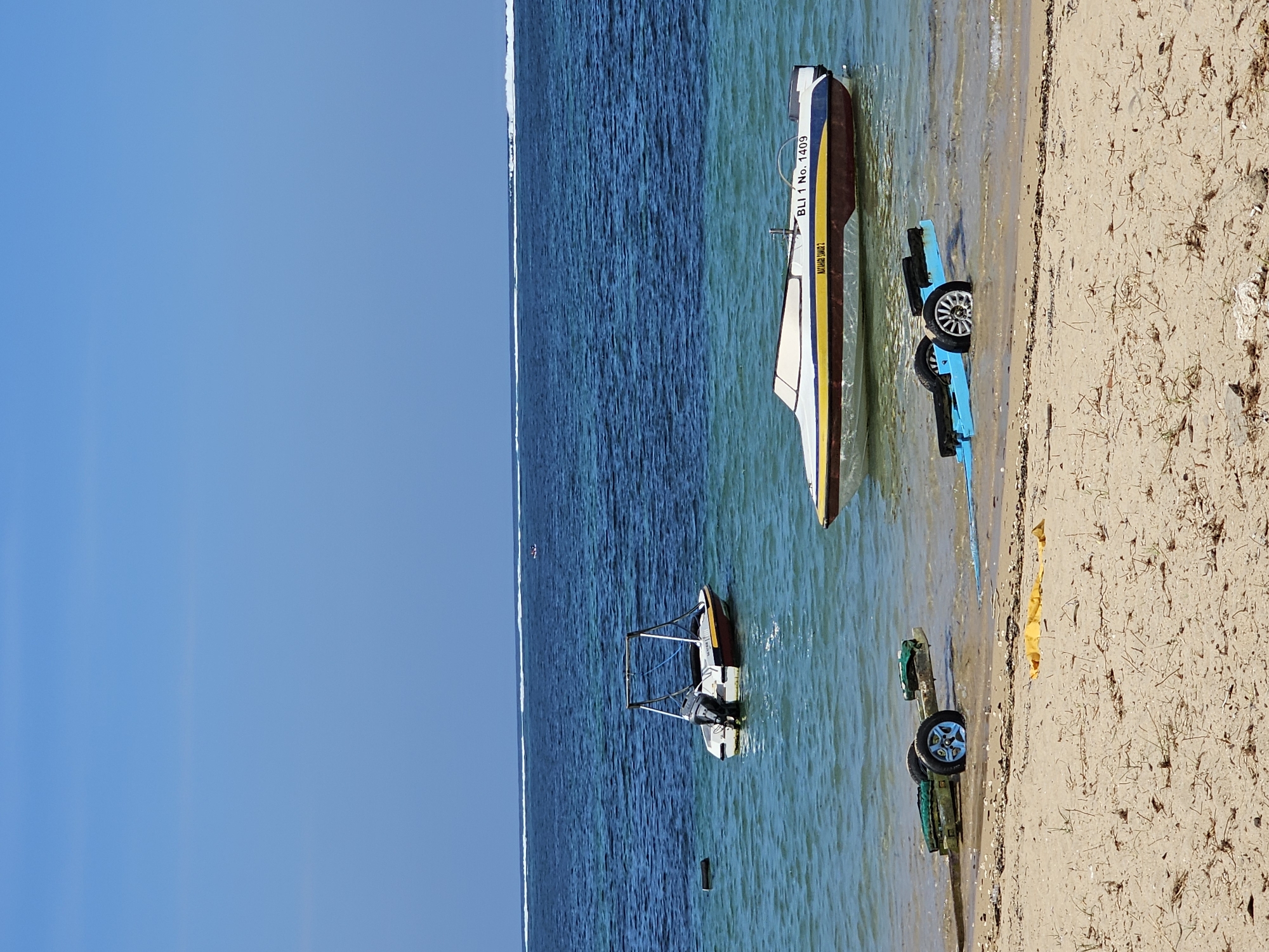 A clear, sunny day at a beach in Denpasar, Indonesia, shows two boats in the turquoise water near the shore. The boat in the foreground is a speed boat with white, blue, and yellow accents, bearing the registration "BLI 1 No. 1409". It is partially submerged in the shallow water. Further out in the water, a smaller boat with a white hull and red trim is also moored. A blue and black boat trailer, with two wheels partially submerged in the water, is positioned on the sandy shore. Another green boat trailer with two blue wheels is visible on the beach to the left. The ocean extends to the horizon, with small waves breaking further out. The sky is a clear, vibrant blue.