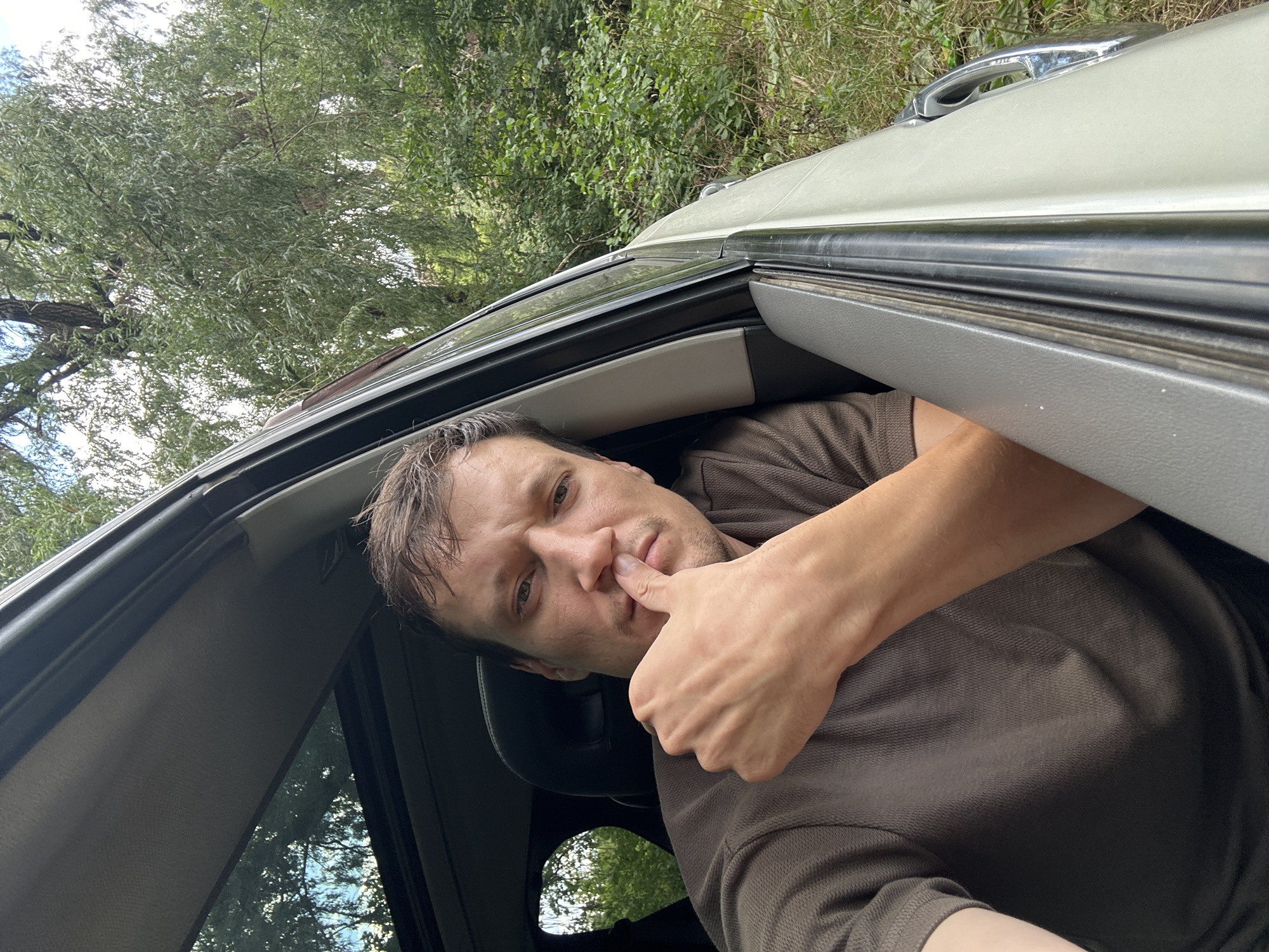 A man is sitting in the driver's seat of a car with the window down, looking into the camera and resting his chin on his thumb. The car is parked outdoors, surrounded by lush green trees and foliage. The man is wearing a brown polo shirt. The sky is visible through the sunroof of the car, reflecting in the window. The scene appears to be in a rural or wooded area.