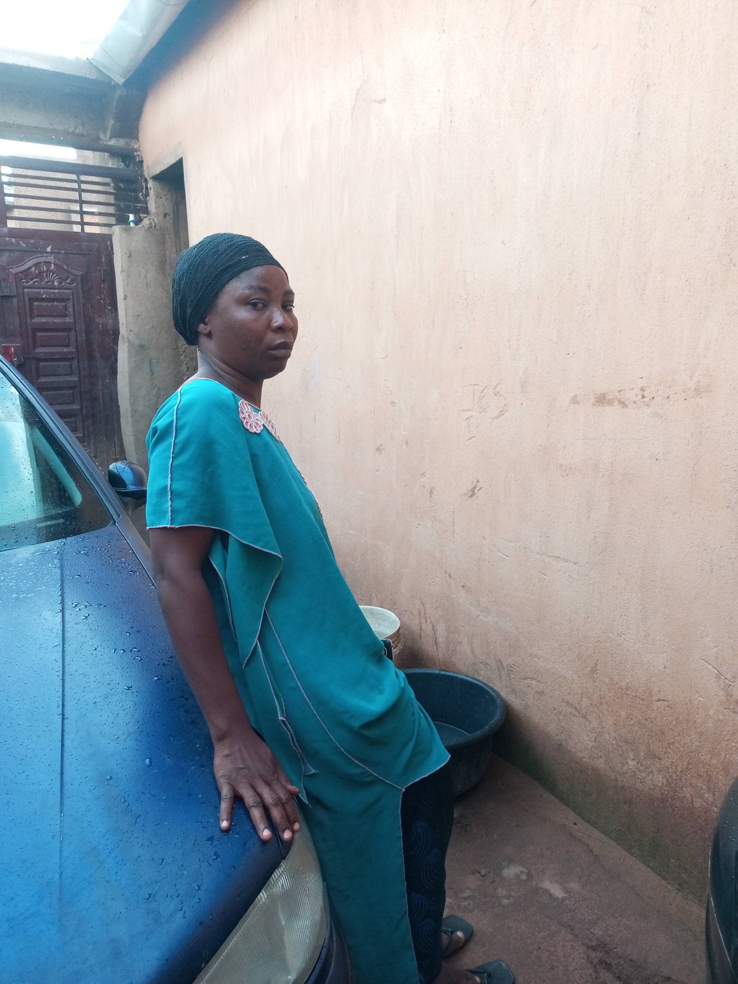 A person wearing a teal tunic and a black headscarf leans against the hood of a wet blue car. The person is standing outdoors, with their right hand resting on the car's hood. In the background, a rough, light-brown wall is visible, with a dark plastic basin and a bucket near its base. To the left of the wall, a portion of a dark-colored door with metal grating is seen. The scene appears to be in a residential or open area within Tempure, Nigeria.
