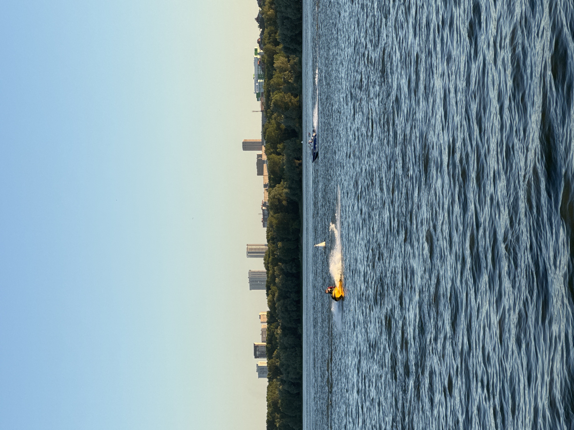 The image depicts a wide, blue river with gentle waves. In the foreground, the water occupies most of the frame. In the middle ground, a yellow jet ski is being ridden by a person wearing a red jacket, creating a spray of water as it moves. Further to the right, a white buoy marks the water. In the background, a dense line of green trees runs along the riverbank. Beyond the trees, a cityscape with several tall buildings is visible against a clear, pale blue sky. The scene suggests a recreational area within or near a city. This is likely the Ob River in Novosibirsk, Russia, given the presence of the river, cityscape, and treeline which are characteristic of the city's waterfront. The time of day appears to be late afternoon or early evening, suggested by the warm, soft lighting.