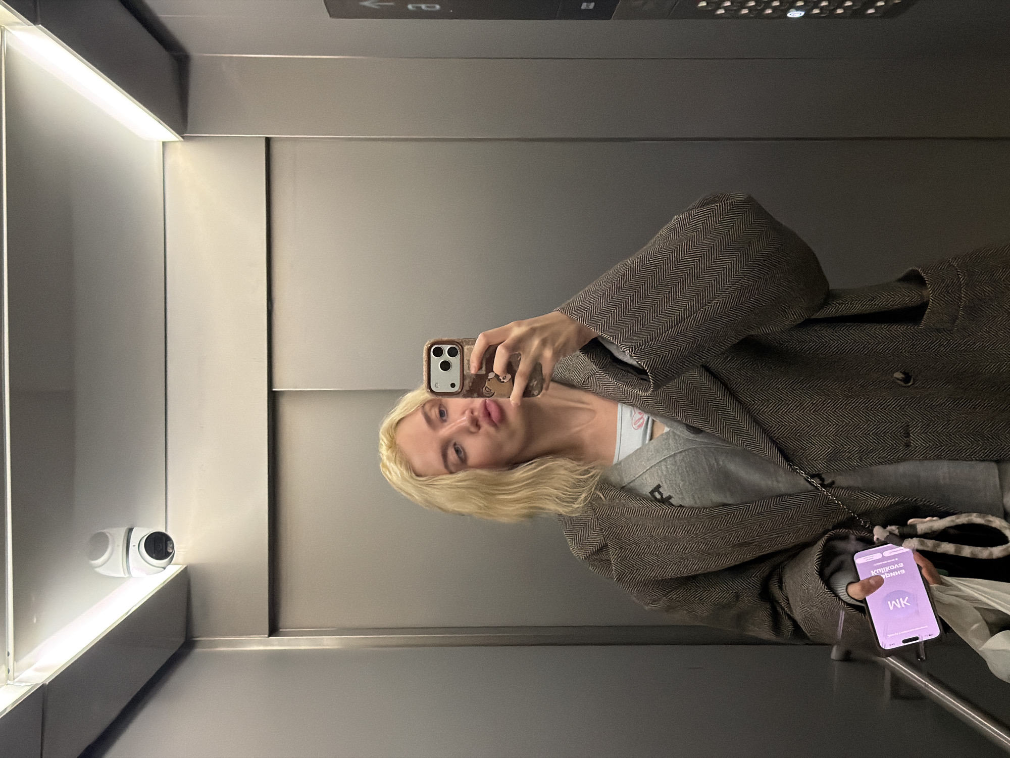 The image displays a young adult female reflected in the polished interior of an elevator, specified as being in Belgrade, Serbia. She has blonde, wavy, shoulder-length hair and is wearing a light-colored crop top, a grey open cardigan, and a dark herringbone patterned overcoat. She holds a smartphone with a brown, bear-themed case in her right hand, angled towards her, indicating she is taking a self-portrait. Her lips are pursed in a prominent pout. In her left hand, she holds a second smartphone with a purple screen displaying "WK" and "SHINGA SYOULD," along with a white plastic bag and a grey and white patterned strap from a separate bag. A white, bullet-style CCTV camera is mounted on the elevator ceiling in the upper left corner, and a strip of bright white light illuminates the ceiling's perimeter. The elevator walls consist of smooth, grey metallic panels, and a section of a dark grey control panel with illuminated buttons and an upward arrow indicator is visible on the right side of the reflection.