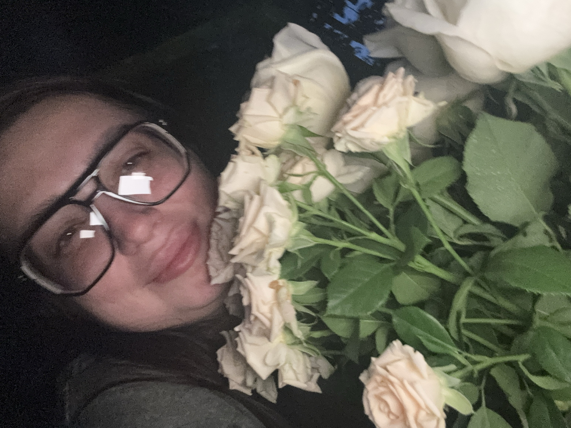 The image displays a close-up of a person wearing glasses, holding a bouquet of light-colored roses. The person's face is partially obscured by the flowers, but they appear to be smiling. The background is dark, suggesting an indoor or nighttime setting. The scene's location in Tolyatti, Russia, is not evident from the visual information provided.