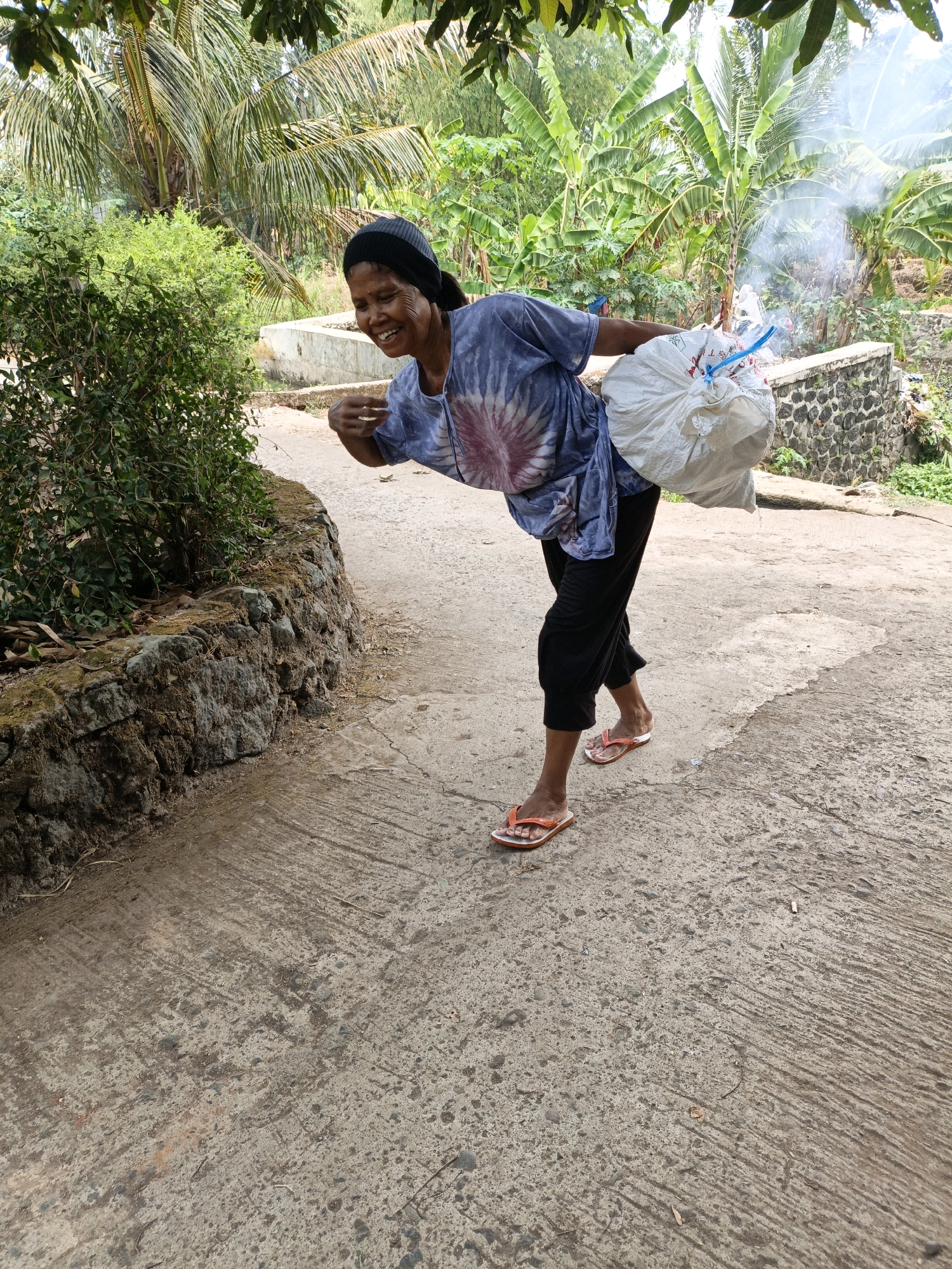 A woman, appearing to be of Indonesian descent, walks on a paved path lined with vegetation and stone walls. She is wearing a tie-dye t-shirt, black pants, and orange flip-flops. The woman carries a large, white plastic bag filled with an unidentifiable substance over her right shoulder. She is smiling and looking to her left. In the background, lush green foliage, including banana trees and palm trees, is visible. The presence of smoke suggests a fire or some industrial activity in the distance. The scene is set in a rural or semi-rural environment, possibly in Mangli, Indonesia, given the vegetation and architecture.