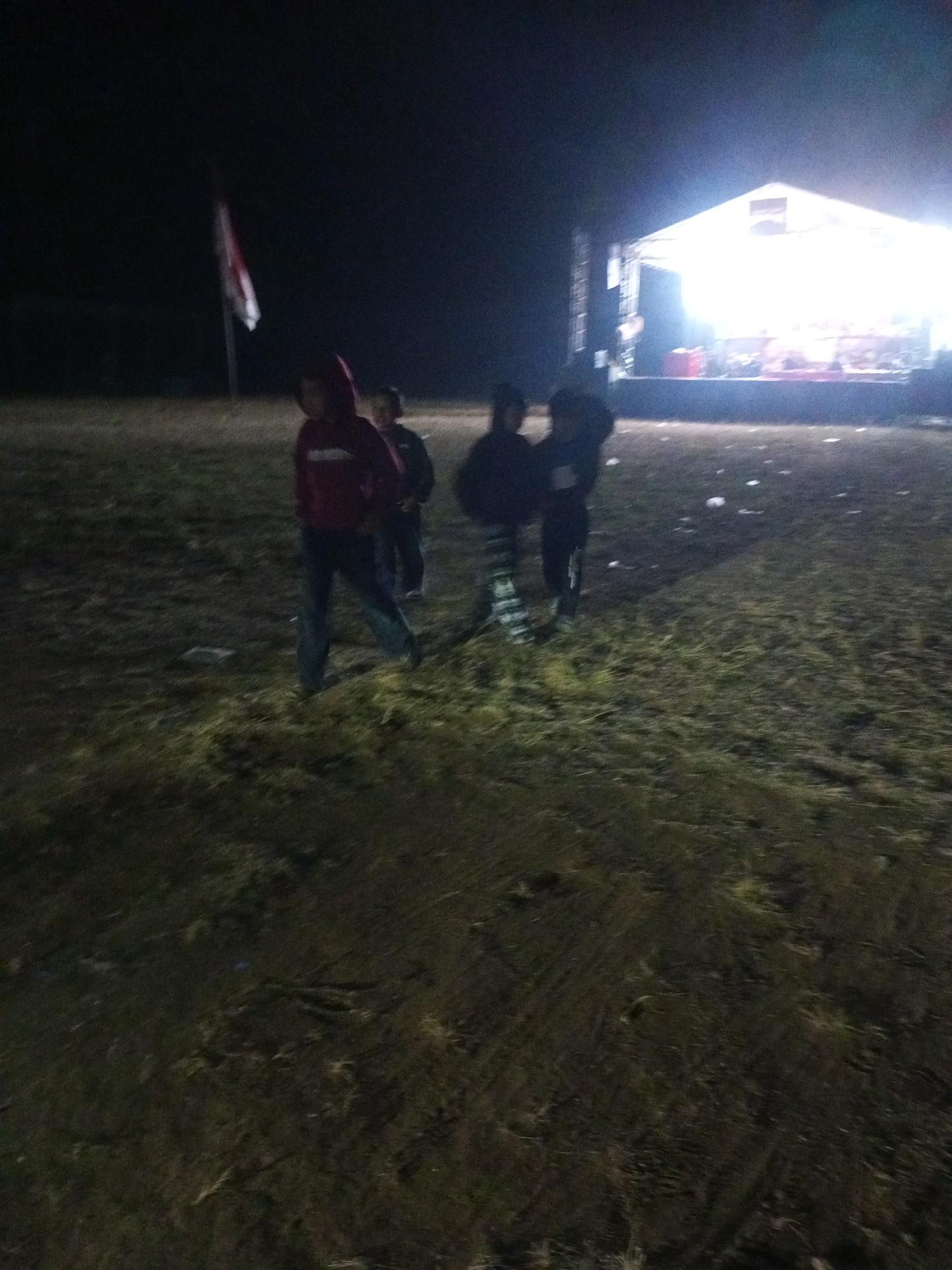 The image depicts a nighttime outdoor scene in the city of Ngadikerso, Indonesia. In the foreground, three individuals are visible, appearing to be children or young teenagers. The person on the left wears a dark red hooded sweatshirt and dark pants, facing away from the camera. To their right, another individual in dark clothing stands with their back to the viewer. A third person, also in dark clothing, is to the right of the second individual, facing partially away. The ground is covered in grass and appears somewhat uneven, with some debris scattered on it.
In the background, a brightly lit stage structure is present, suggesting an event or performance is taking place. A flag, appearing to be red and white, is mounted on a pole to the left of the stage, partially obscured by darkness. The lighting from the stage illuminates the immediate area around it. The overall environment appears to be an open field or park.