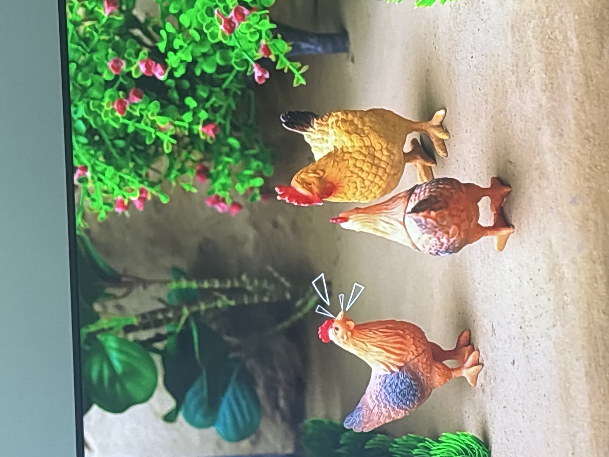 This image depicts a close-up, eye-level view of three miniature plastic chickens arranged in what appears to be a diorama. The chickens are positioned on a sandy-colored surface, with artificial greenery and a plant with pink blossoms forming the background. One chicken, facing left, has a red comb and wattle, and its beak is open, with white triangular shapes emanating from it, suggesting it is clucking or vocalizing. It has orange and brown plumage on its body and blue feathers on its back. Another chicken, facing away from the viewer, is mostly brown with red accents and is standing slightly to the right of the first chicken. The third chicken, larger and facing right, is a bright yellow and orange color with a prominent red comb and wattle. It is positioned behind and to the right of the other two chickens. The overall scene suggests a playful or decorative arrangement. There is no indication of the city of Dubai, United Arab Emirates, or any forensic context.