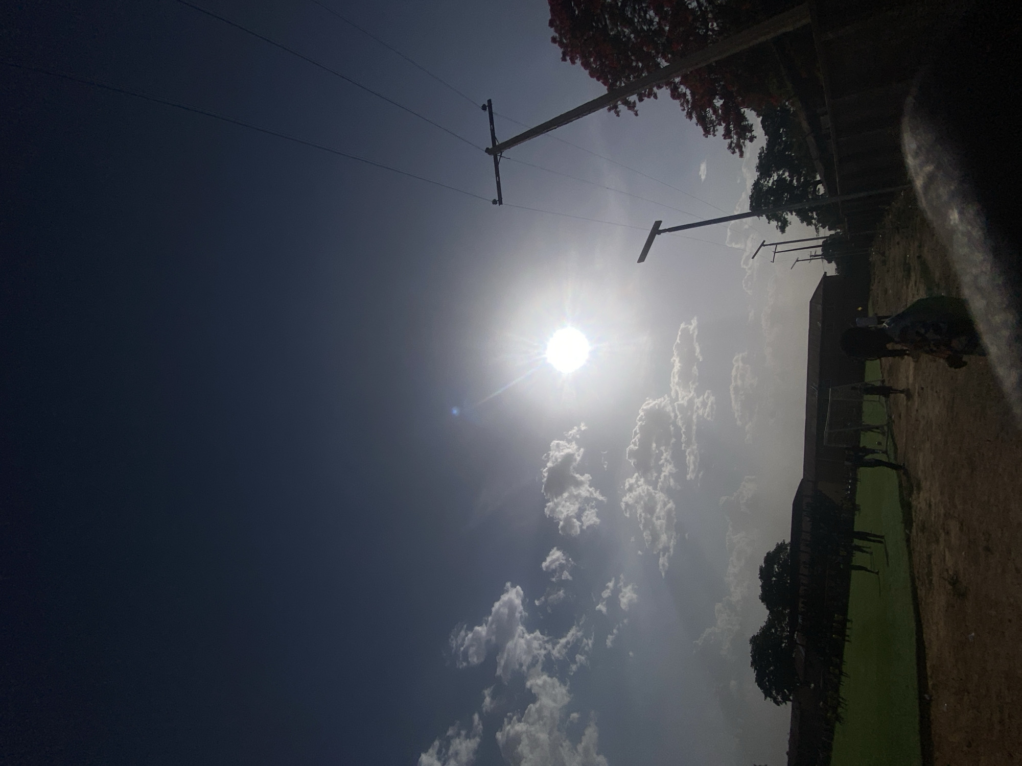 The image depicts a bright, sunlit scene in Tudun Wada, Nigeria. Overhead power lines and utility poles are visible. Clouds are scattered across the sky, which transitions from dark blue to a lighter shade near the horizon. A sports field borders a building, with figures appearing to be engaged in activity on the field. Trees line the perimeter of the field. Part of the camera's operator (perhaps a finger) is visible on the right side of the image.