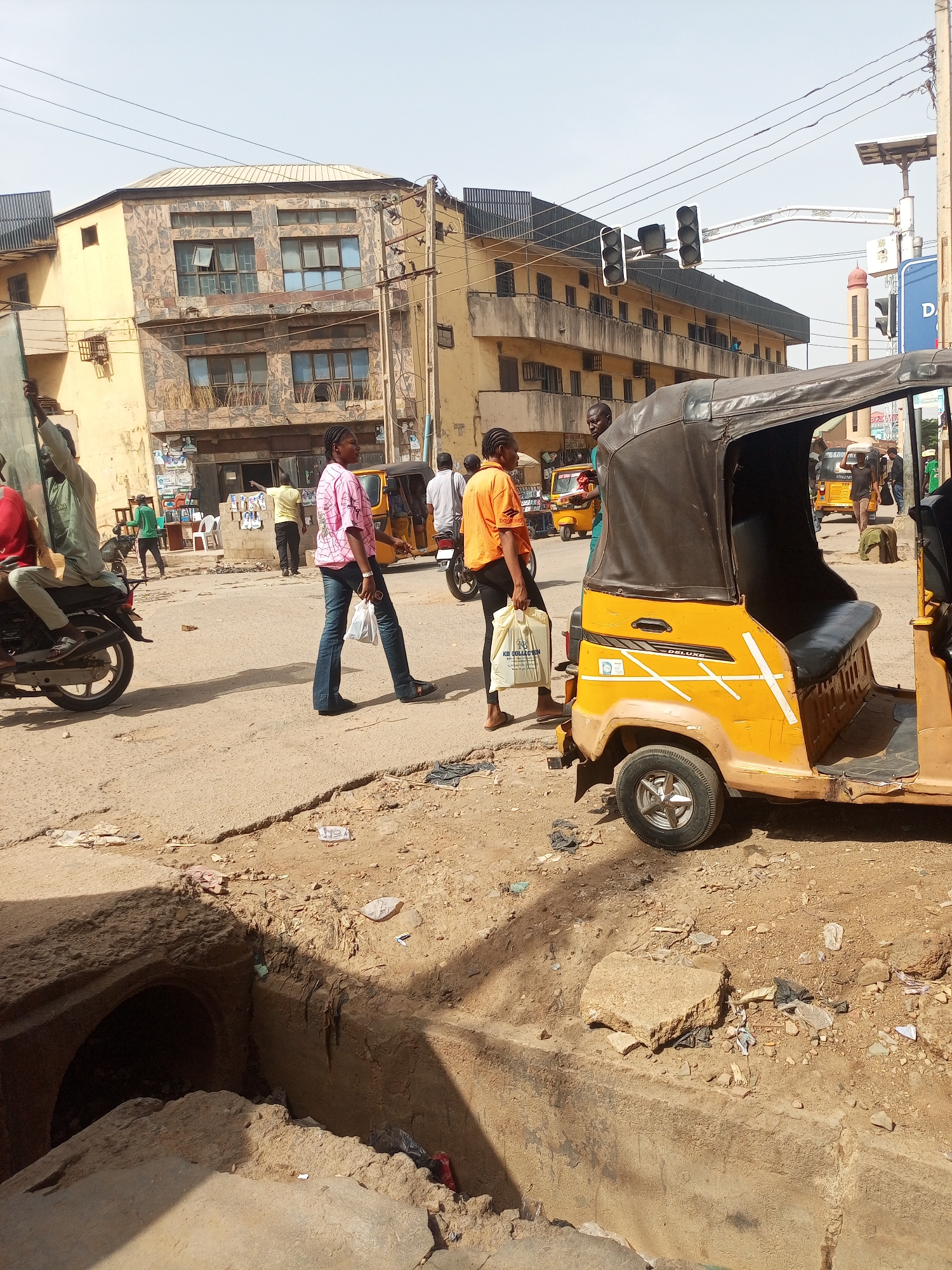 The image captures an urban scene in Maiburiji, Nigeria. The foreground shows an open drainage ditch alongside a dilapidated, unpaved road. Litter and debris are scattered across the road surface and in the ditch.
A yellow "Keke Napep" (auto rickshaw) is partially visible on the right side of the frame. Two people are walking past the auto rickshaw; one in a pink patterned shirt and jeans, and the other in an orange shirt carrying a white shopping bag. Another Keke Napep is seen further down the street.
In the background, several buildings are visible. One is a yellow three-story building with some sort of tiling pattern on a part of the facade. Another building behind it is visible with a darker roof. A traffic light is seen overhead.
Other individuals are seen in the background, either walking or standing near buildings. A motorcycle is parked on the left. Overall, the scene portrays a busy, somewhat run-down urban environment.