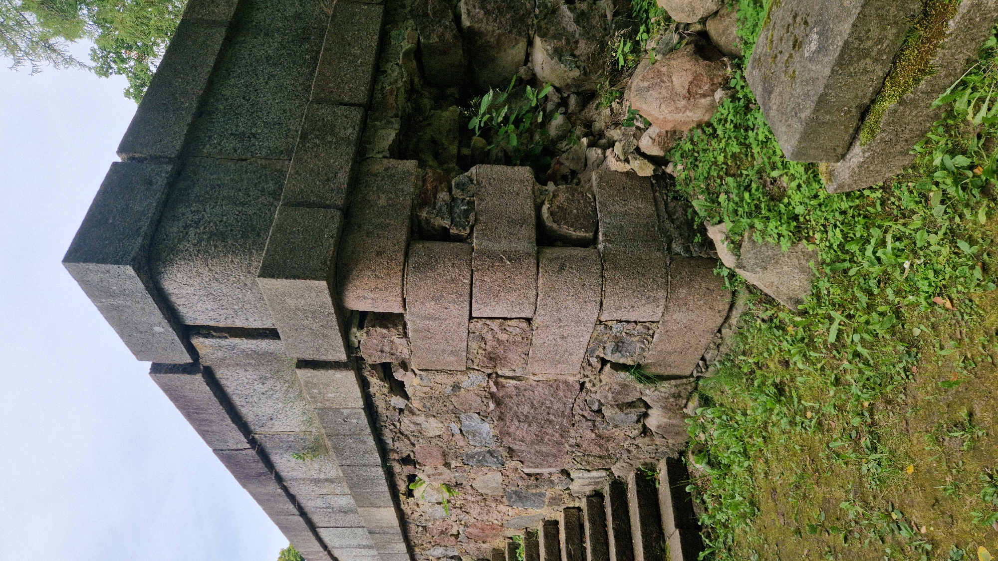 An outdoor, low-angle, eye-level medium shot captures a section of a dilapidated stone structure, likely part of the historical Kazdanga Manor ruins in Kazdanga, Latvia. The structure exhibits a corner where a lower, roughly constructed stone wall meets a more precisely engineered upper section made of large, rectangular granite blocks. Some of these upper blocks show signs of weathering and displacement, with noticeable gaps and cracks between them. The lower wall is composed of irregularly shaped stones of varying sizes and colors, embedded with mortar.
To the left of the structure, a set of stone steps, also appearing weathered, ascends alongside the wall. The ground surrounding the structure is covered in a mixture of moss, grass, and small green plants, with several large, rounded stones and broken pieces of masonry scattered about. In the background, the upper parts of the stone construction are visible, extending upwards and disappearing out of frame. Lush green foliage and trees are visible at the top right of the image, suggesting a natural, overgrown environment. The sky above is overcast and pale. No people or animals are present in the scene.